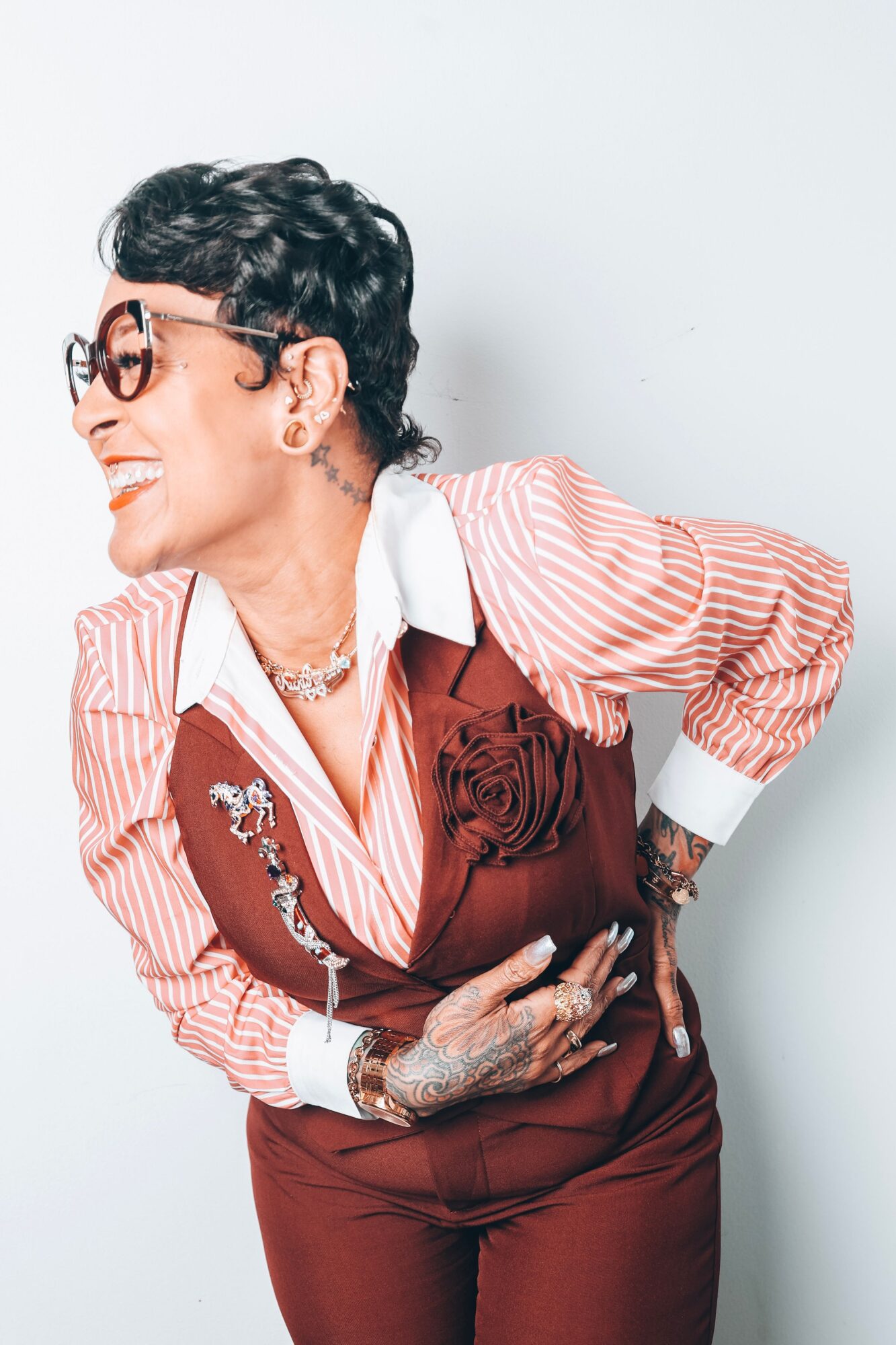 Woman with short dark hair, glasses, smiling, wearing a striped shirt and a vest with a large flower, standing against a light background.