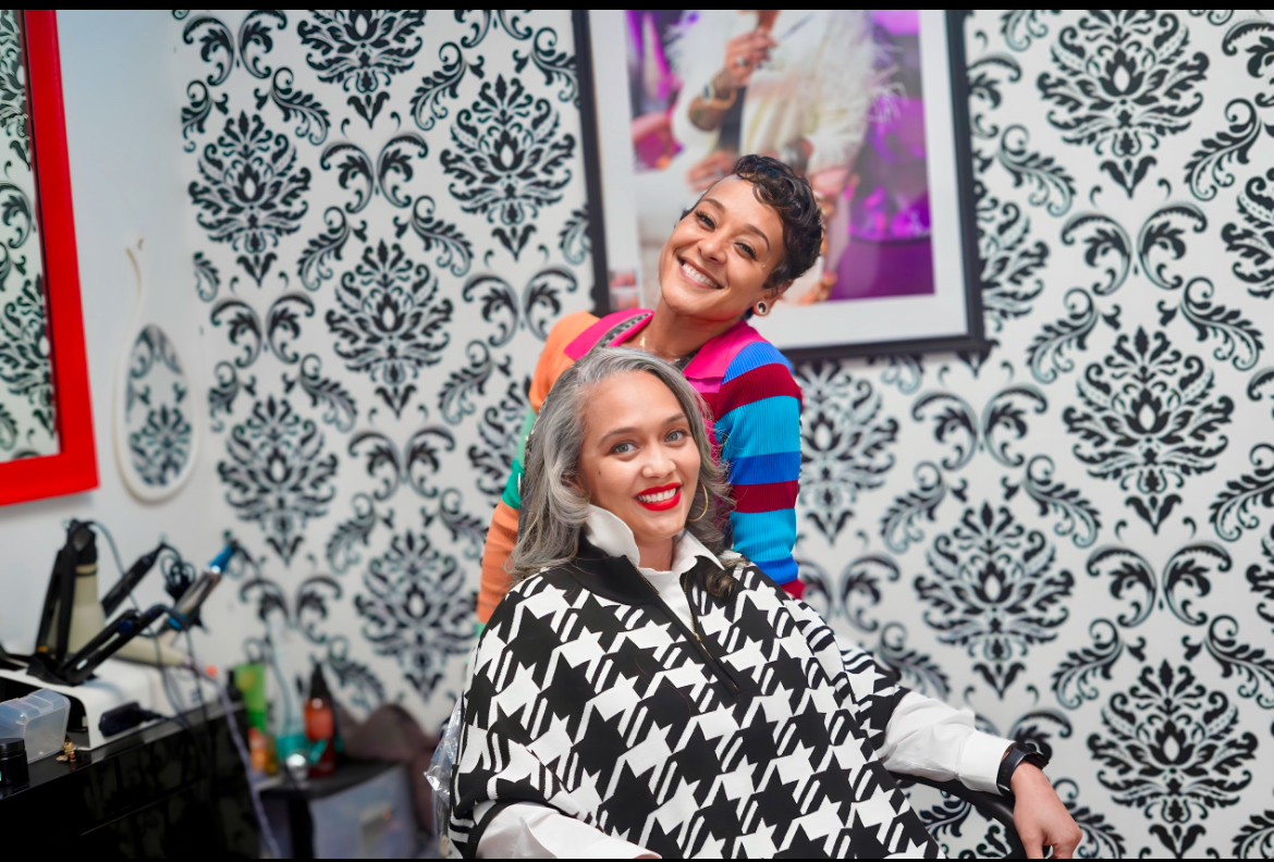 Two women smiling in a salon, one sitting with a cape, the other standing behind her, patterned wallpaper in background.