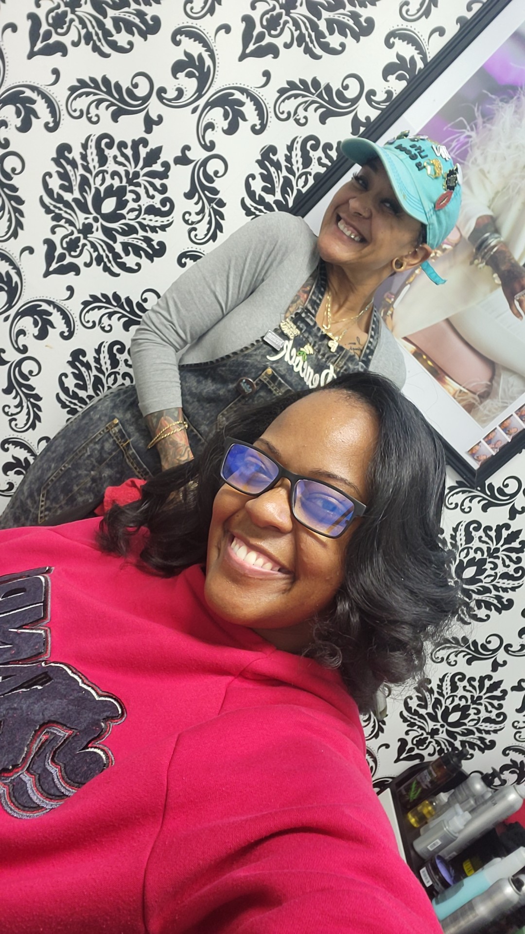 Two women smiling, one wearing glasses and a red top, the other wearing a hat and gray cardigan, in front of patterned wall.