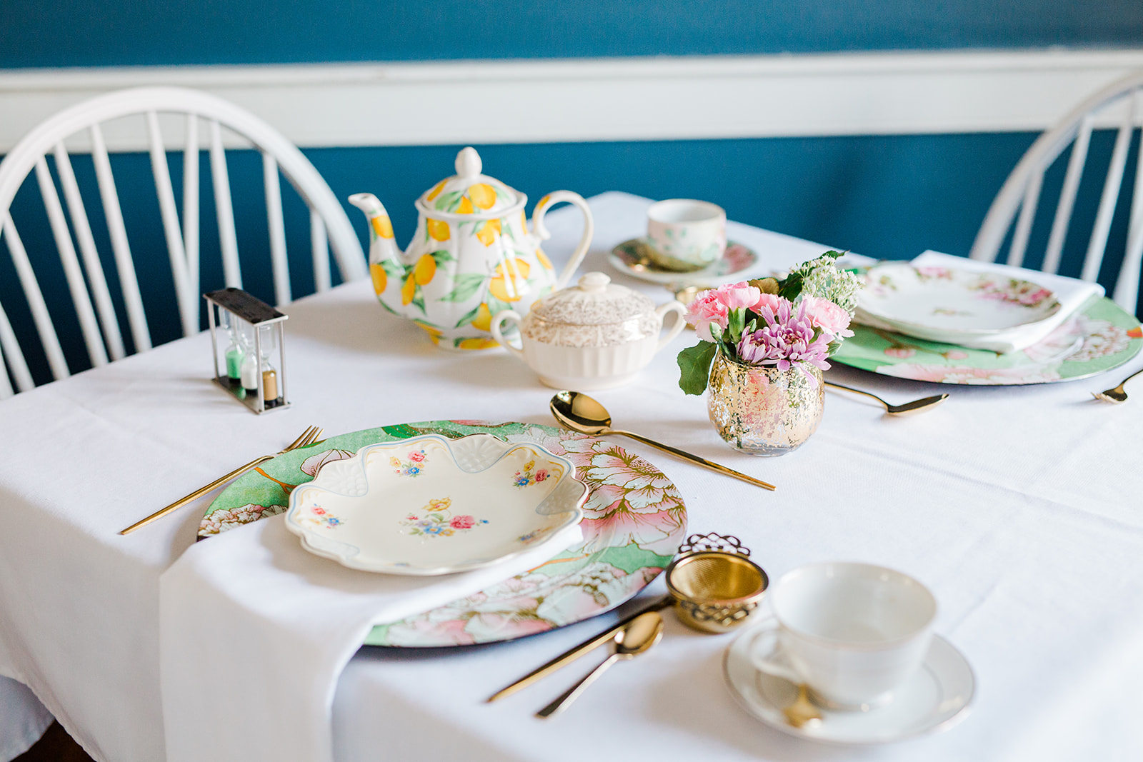 Table set with teapot, cups, plates, flowers, and utensils, with chairs in the background.