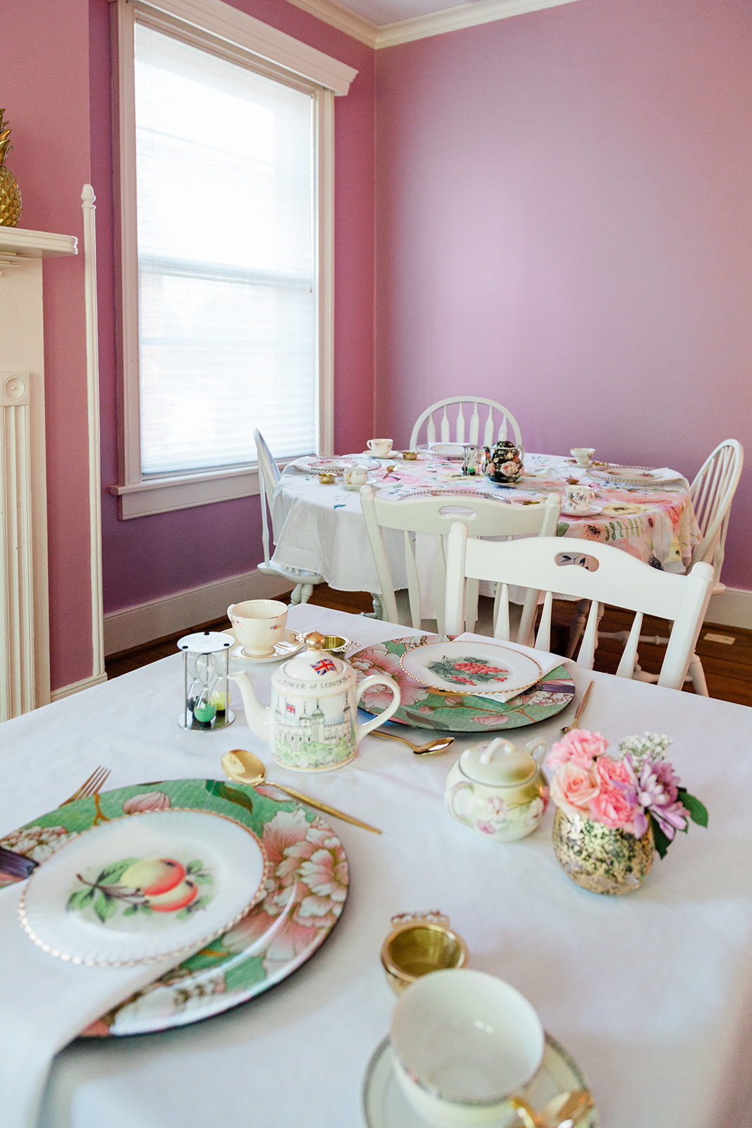 Dining room with pink walls, white window frame, and a table set with plates, teapot, and flowers.