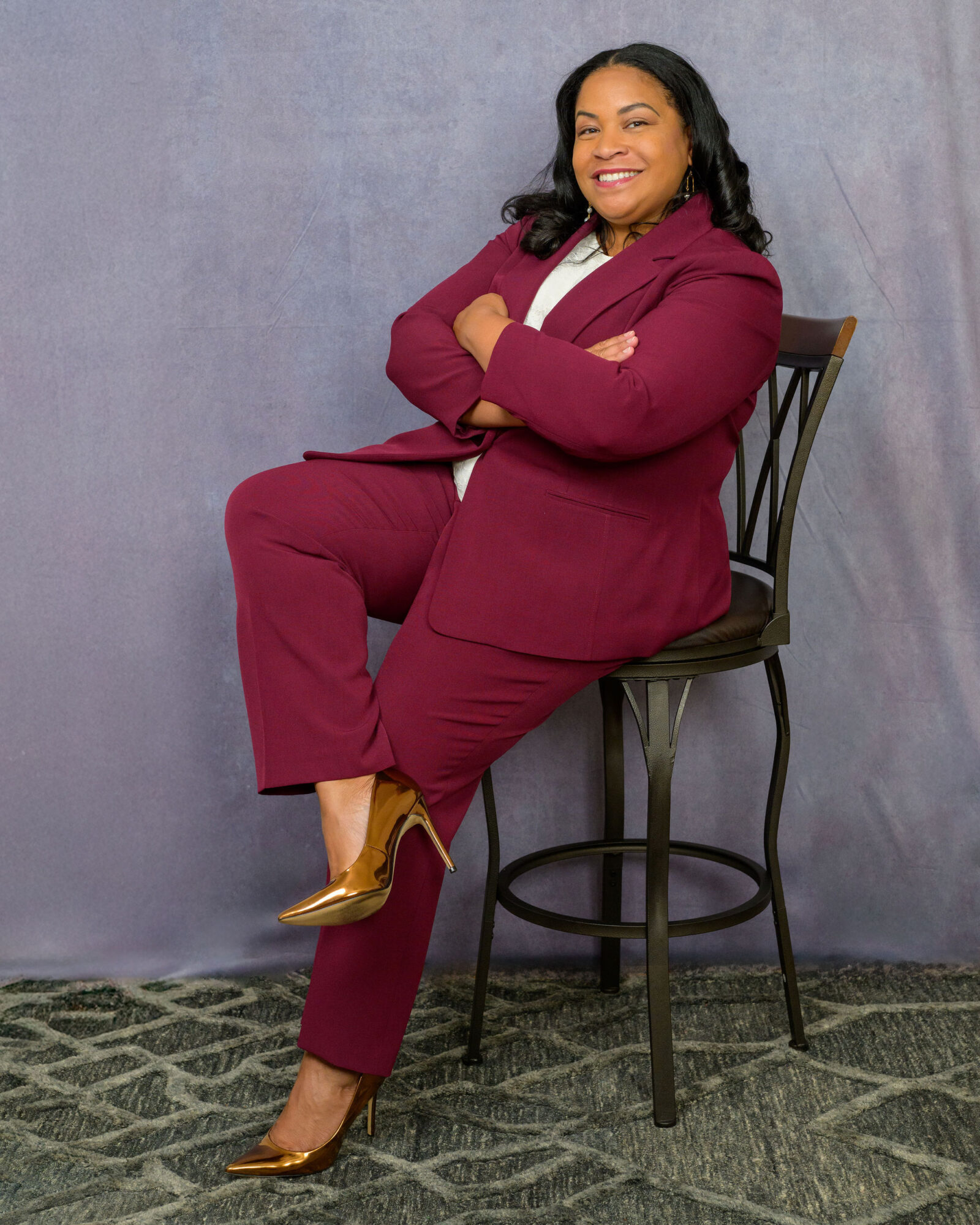 Woman in a maroon suit sitting on a high chair with arms crossed, smiling, against a plain background.