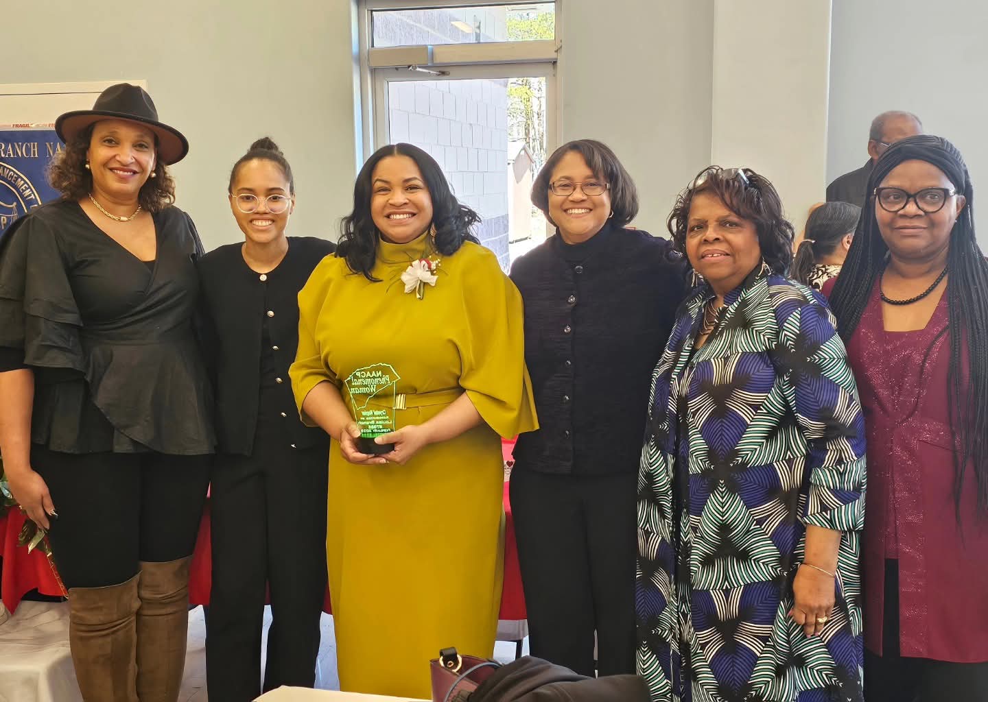 Group of six women standing indoors, smiling, one holding an award, with a window behind them.