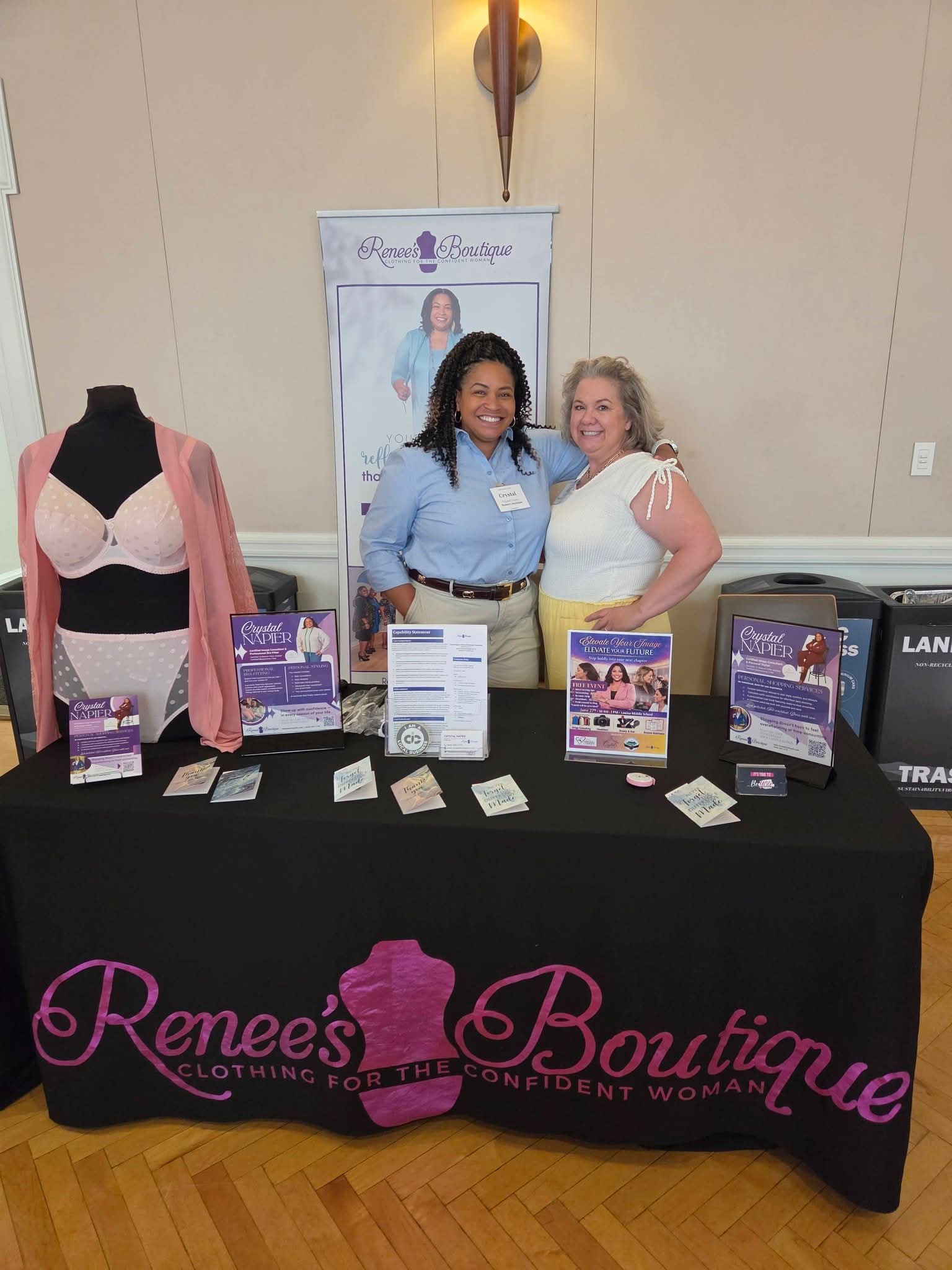 Two women stand behind a table with promotional materials, in front of a banner and a mannequin with clothing, in a room with wooden floor.