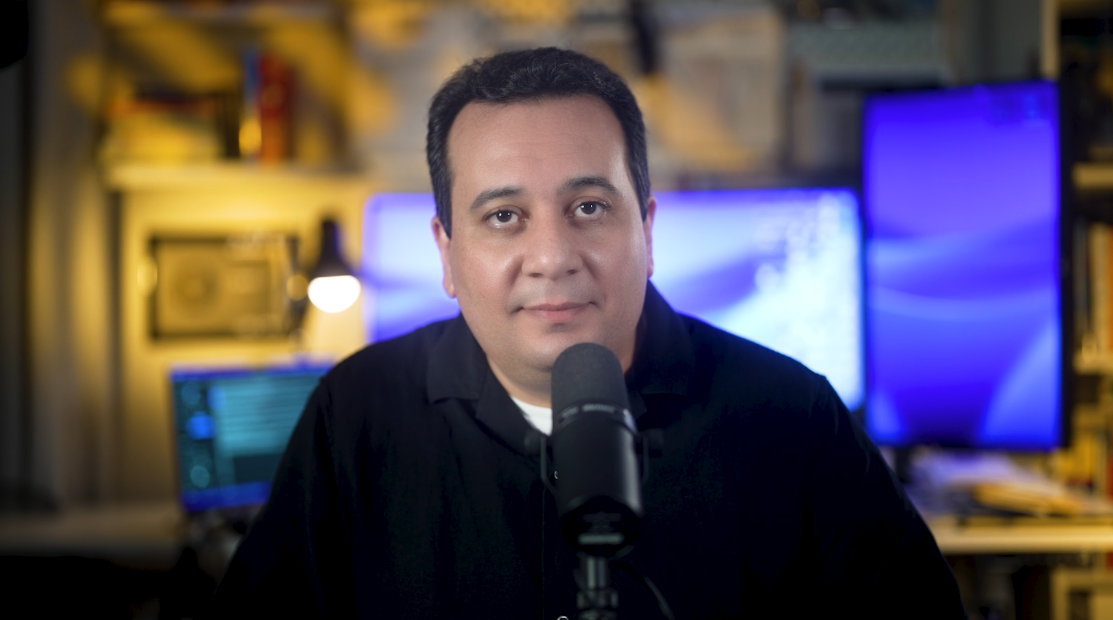 Man with dark hair and a black shirt speaking into a microphone in a studio with monitors and equipment behind him.