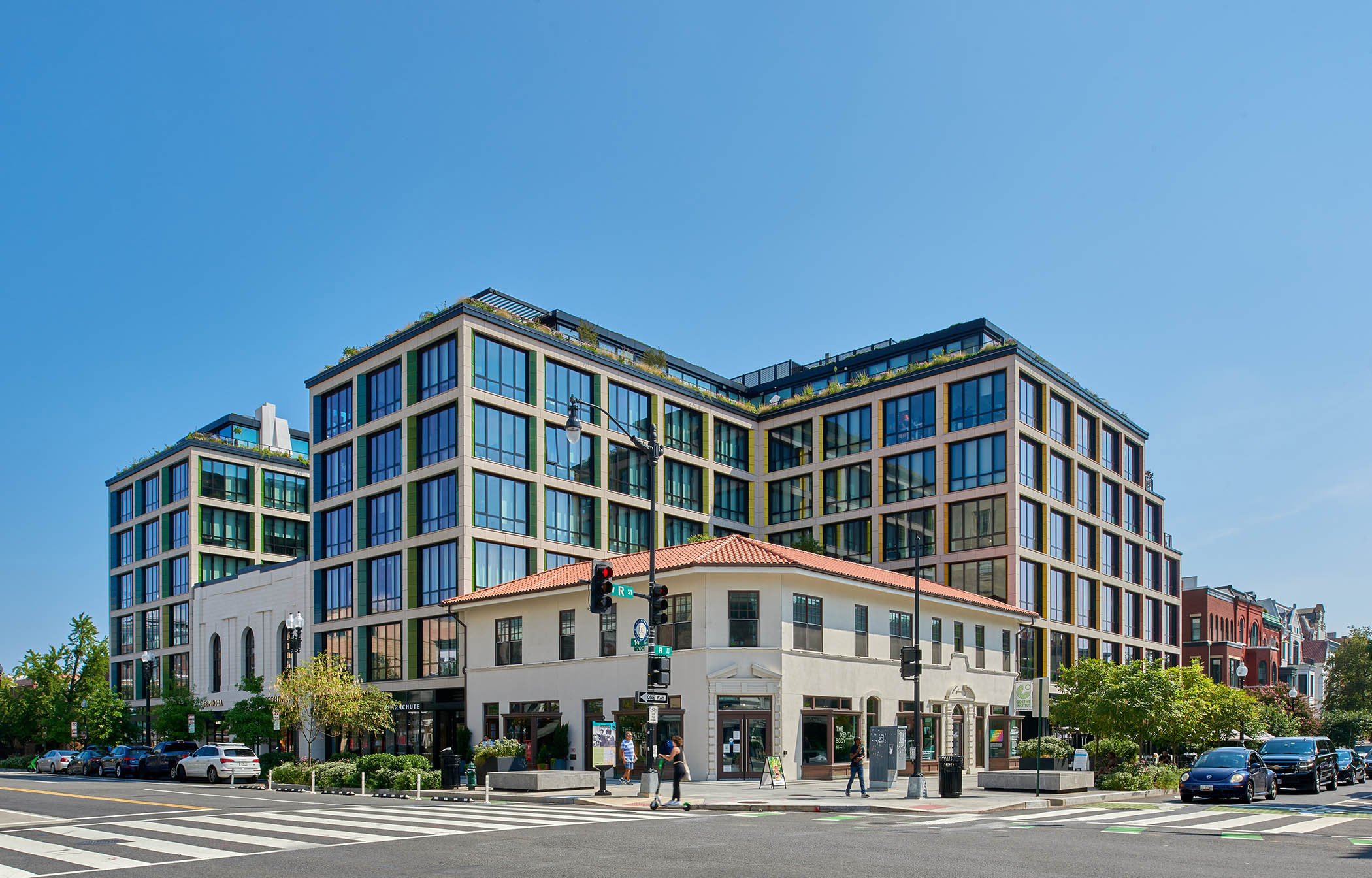 Modern multi-story building with large windows at a busy street corner, pedestrians, and cars, under a clear blue sky.