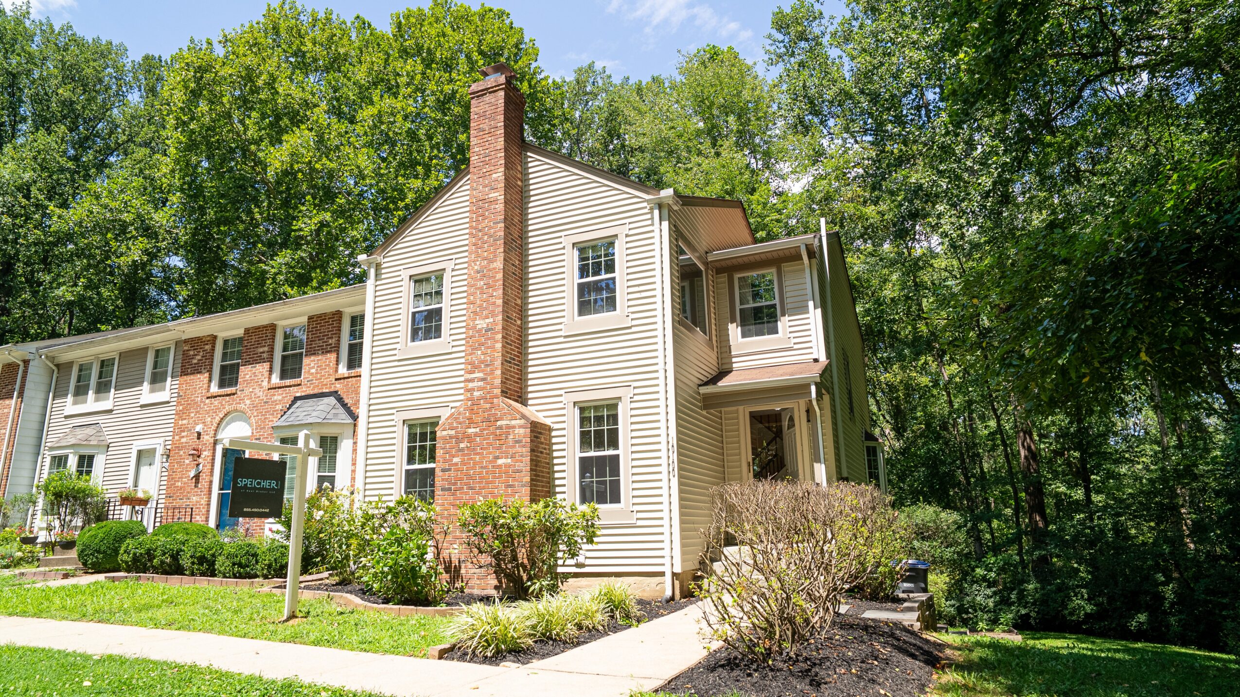 Multi-story house with brick chimney, surrounded by green trees and landscaped yard, under a blue sky.