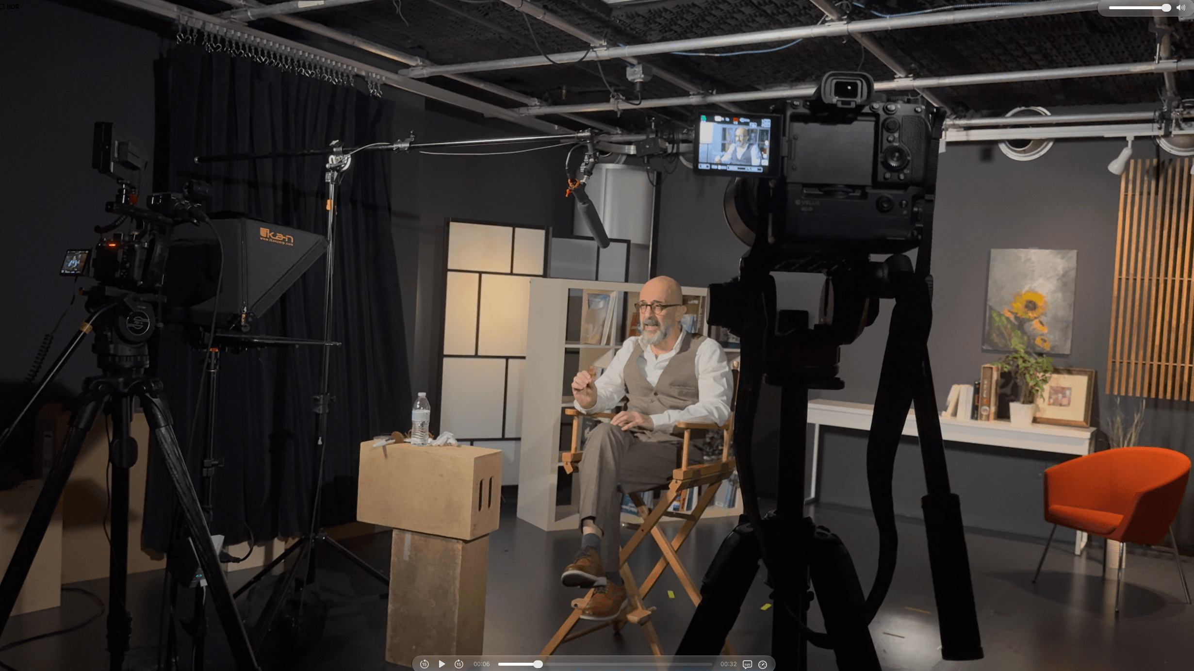 Person sitting on a chair in a studio with cameras and lighting equipment, a table, and a sofa in the background.