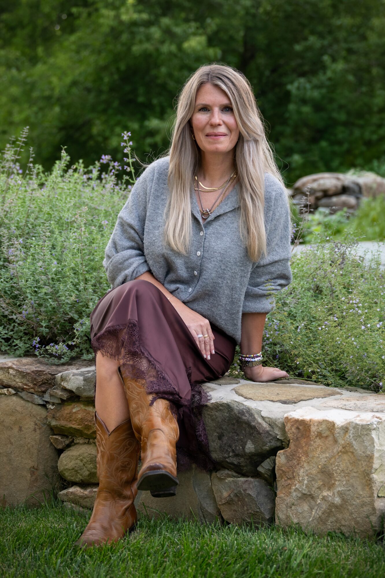 Woman with long blonde hair sitting on a stone wall outdoors, wearing a gray sweater and brown boots, surrounded by greenery.
