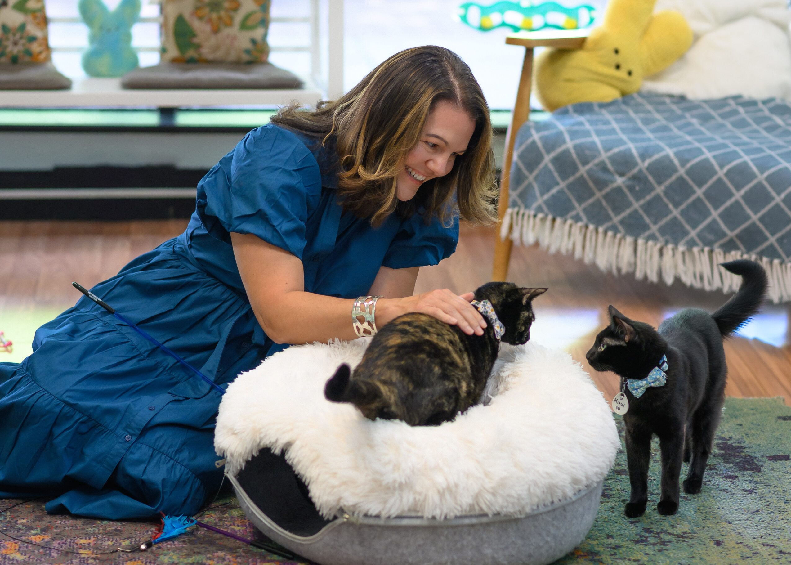 Woman in blue dress petting two cats on a round bed, in a cozy room with a bed and decorative items