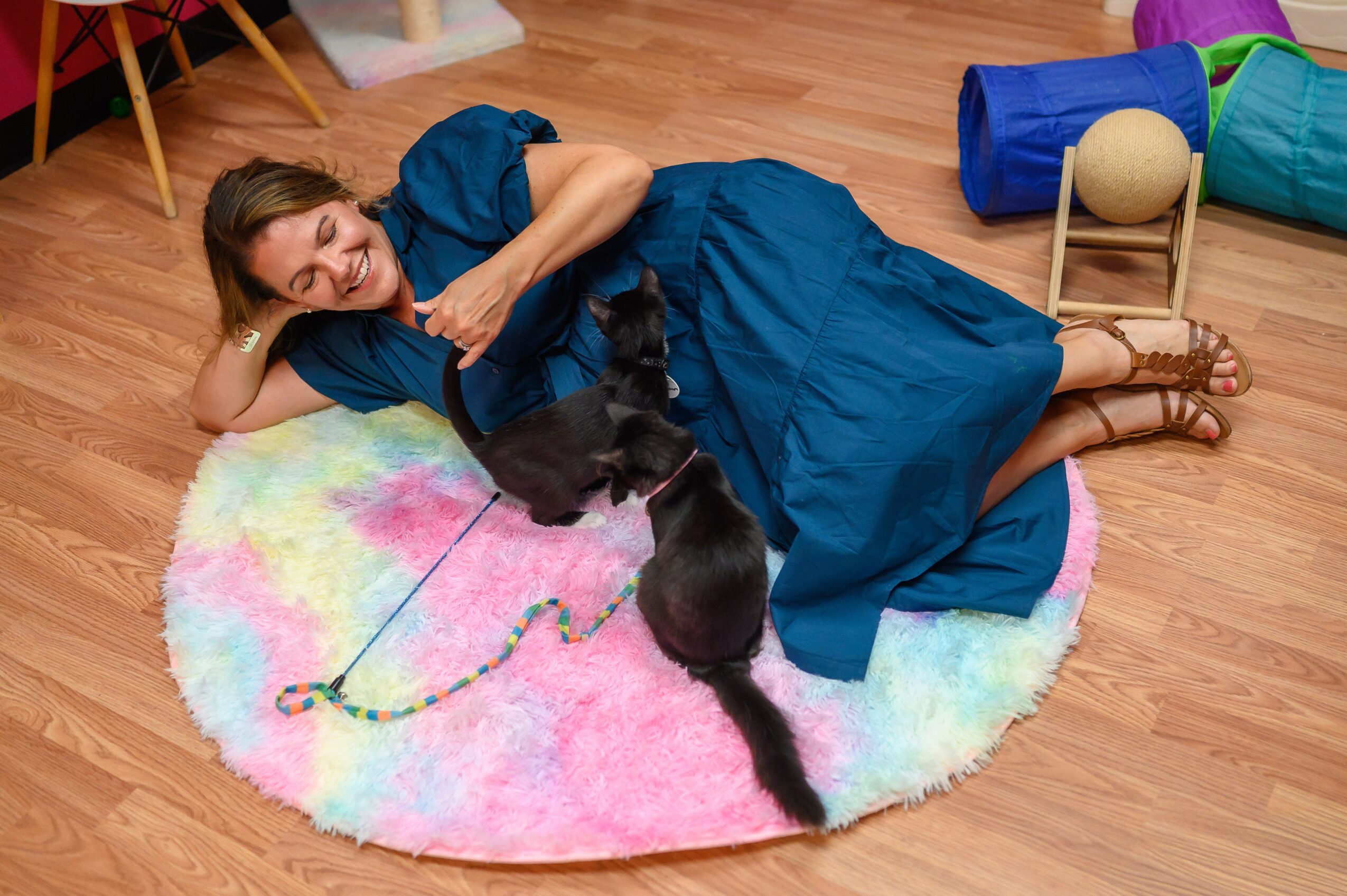 Woman lying on colorful round rug with three black kittens, wooden floor, and toys nearby.