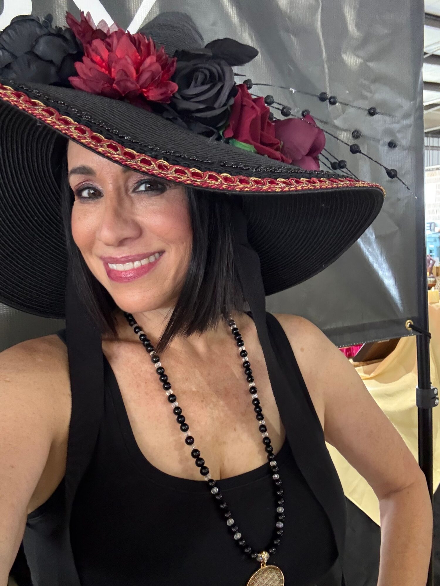 Woman wearing a large black hat with red flowers, smiling, with black clothing and jewelry, in front of a gray background.