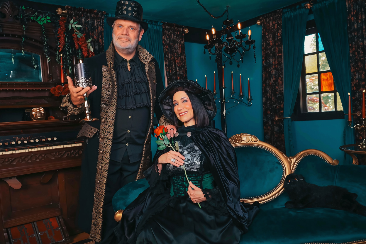 A man and woman dressed in vintage clothing in a dimly lit, ornate room with dark blue walls and antique furniture.
