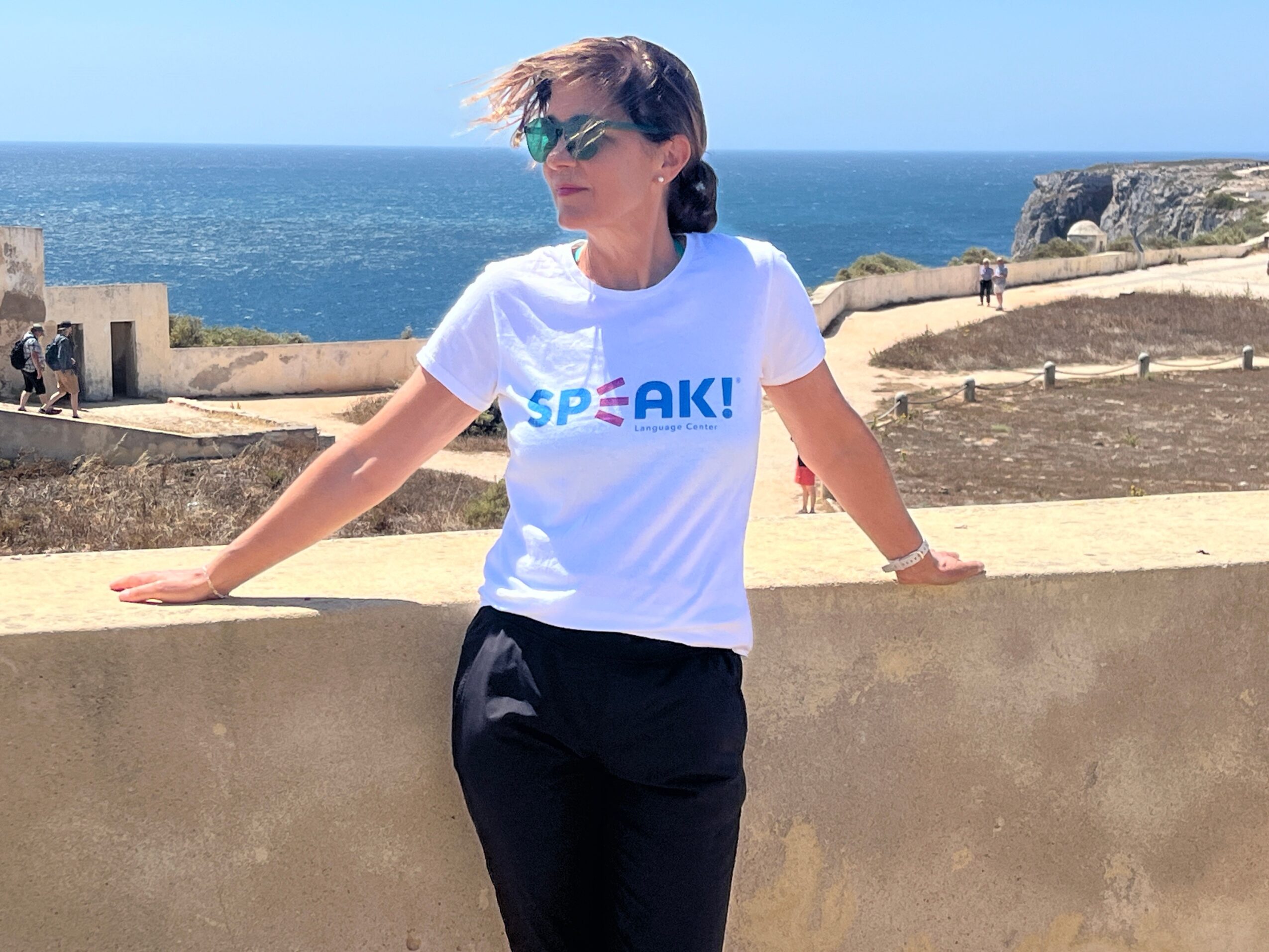 Woman leaning on a wall near the sea, wearing sunglasses and a white T-shirt, with a clear blue sky and ocean in background.