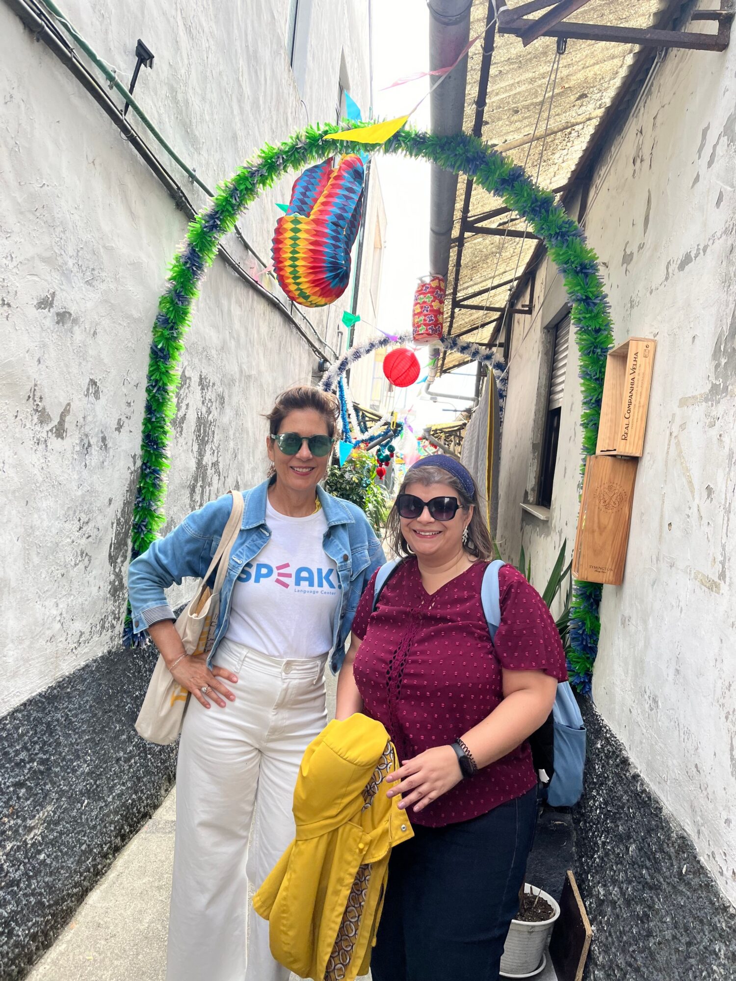 Two women standing outdoors near a colorful heart-shaped decoration, one holding a bouquet of flowers, both smiling, with a building and trees in the background.