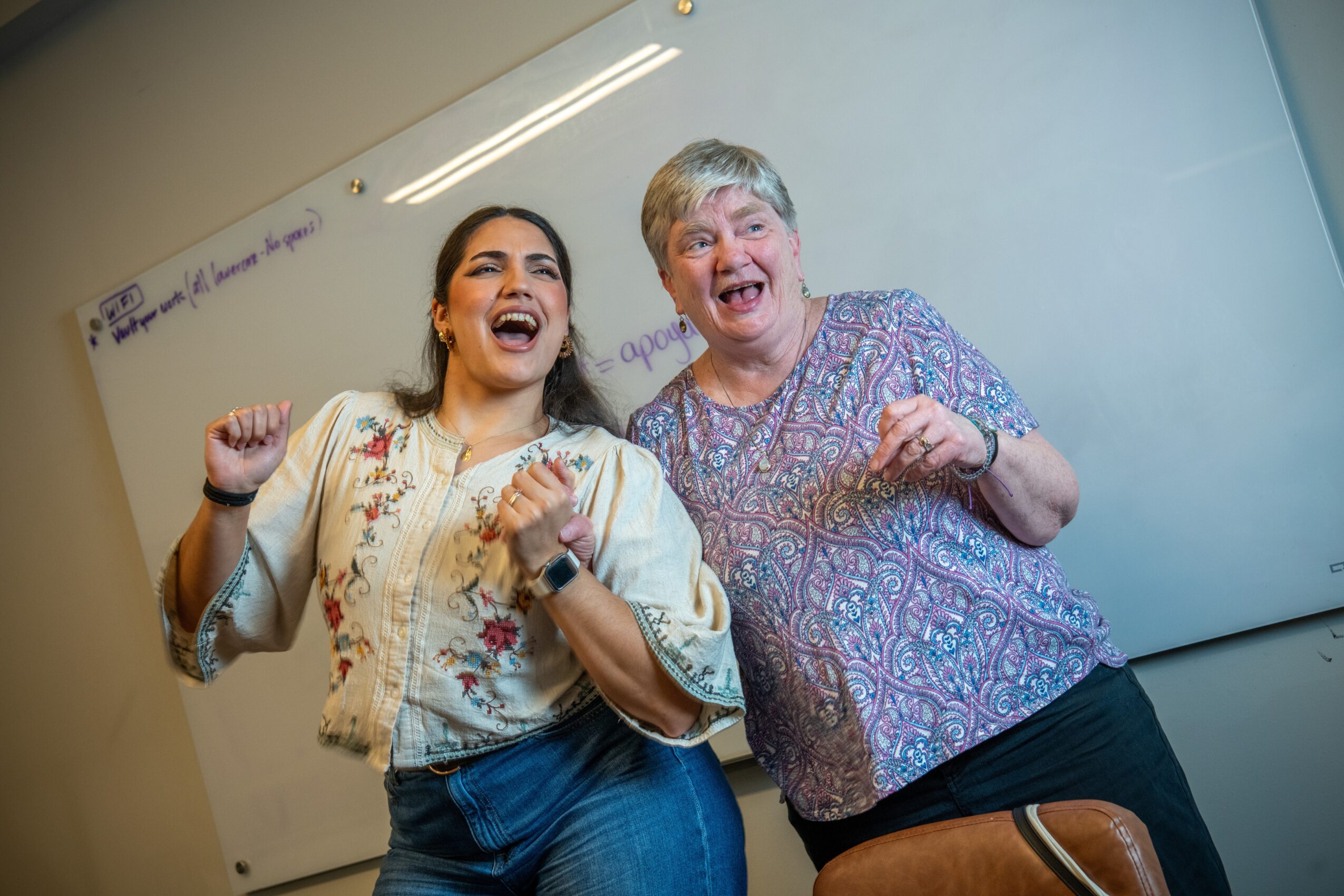 Two women laughing and smiling, standing in front of a whiteboard in a classroom or meeting room.