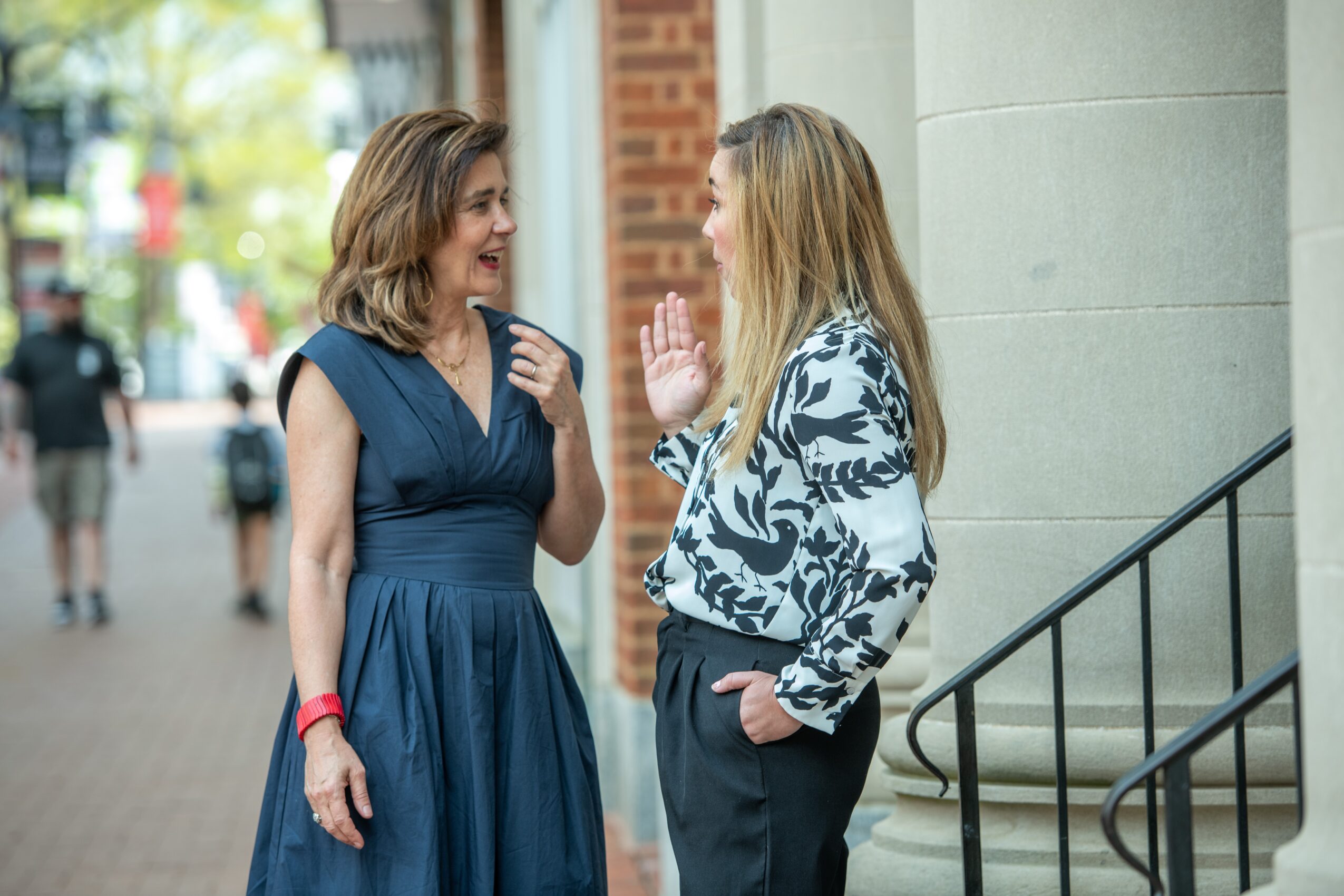Two women are talking outdoors near a building with columns, one gesturing with her hand, the other smiling.