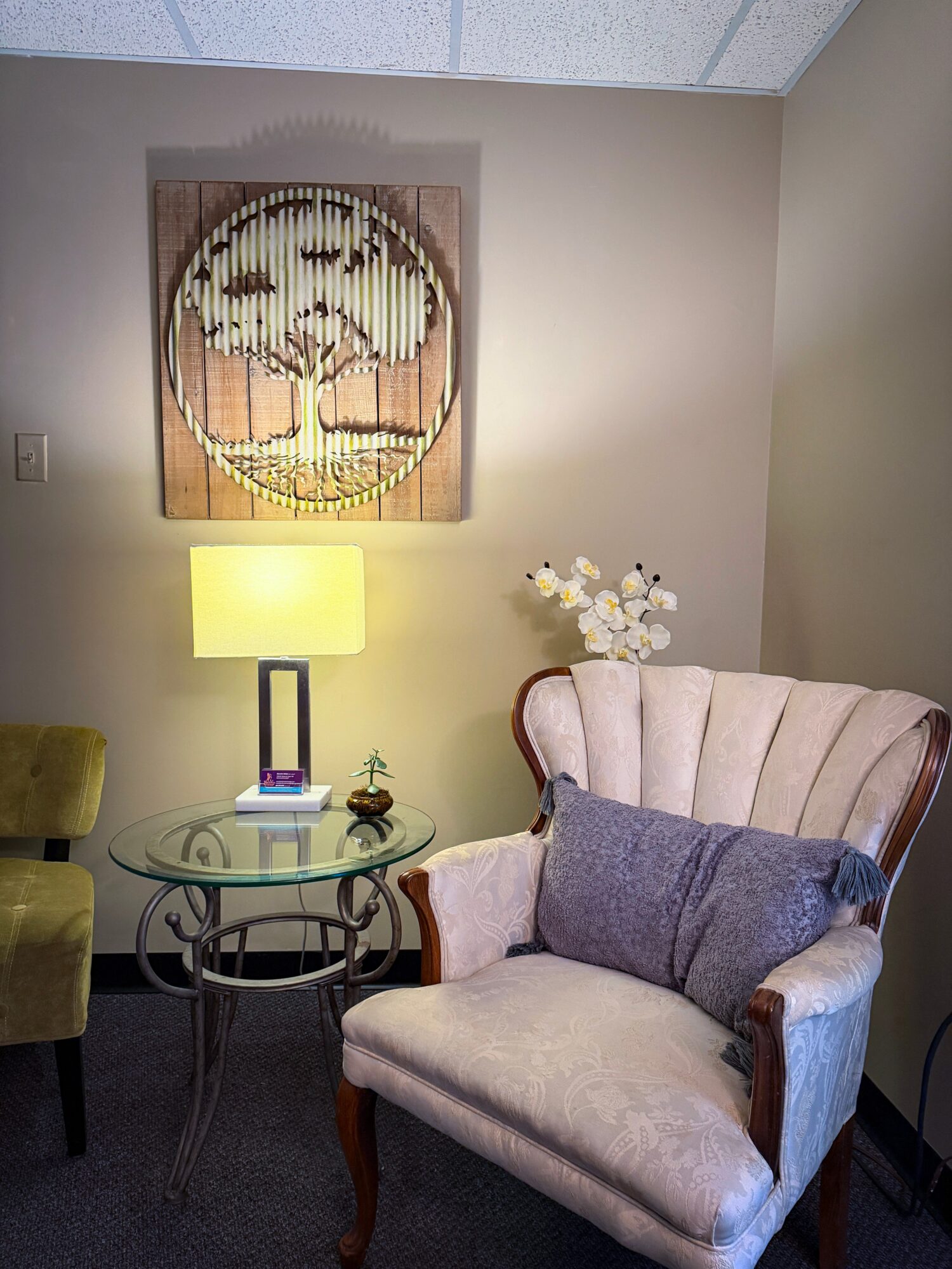 Living room corner with a beige armchair, purple pillow, glass side table, lamp, and wall art, with a flower arrangement.