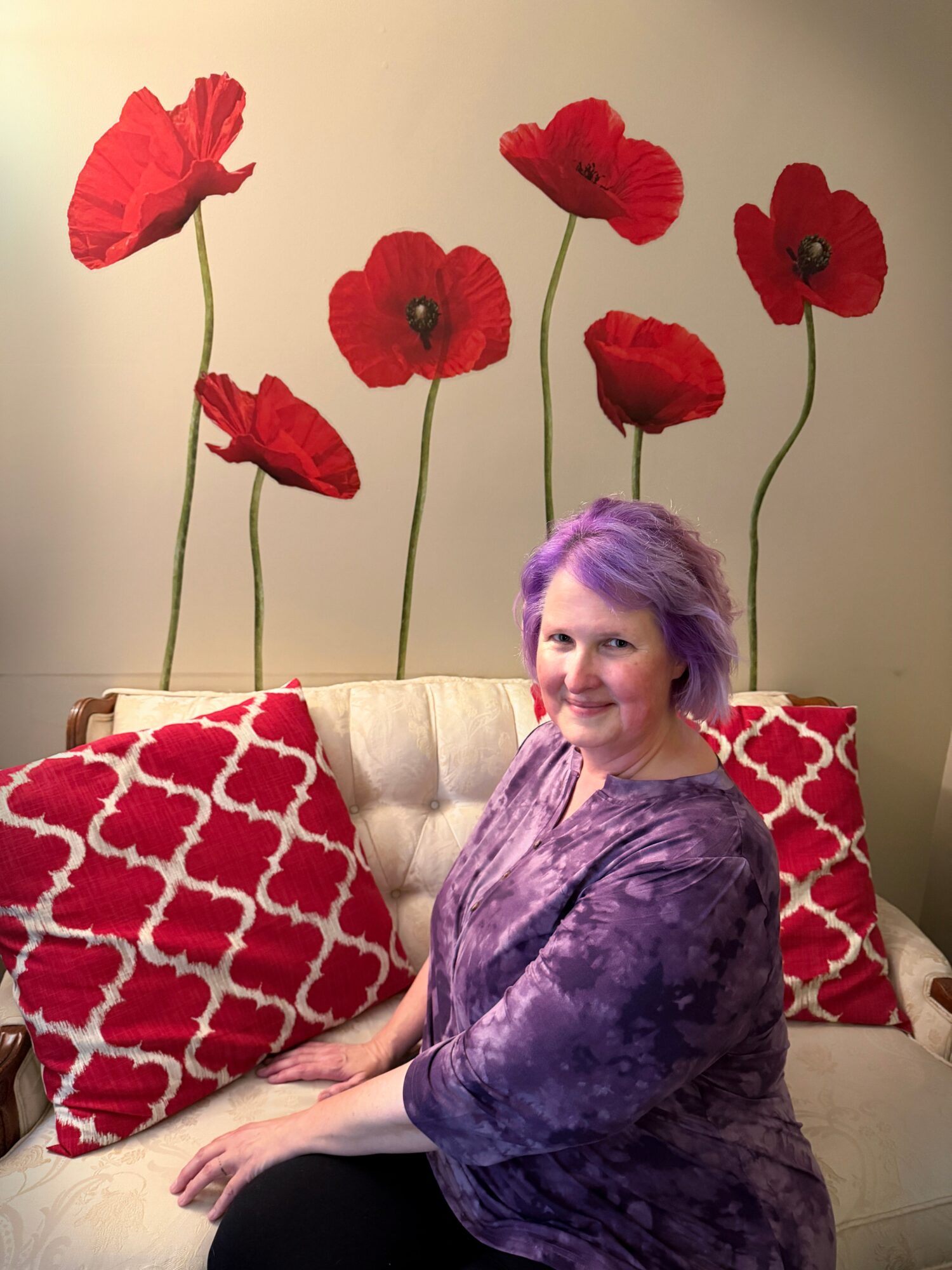 Woman with purple hair sitting on a beige sofa with red patterned pillows, behind large red poppy flower wall art.