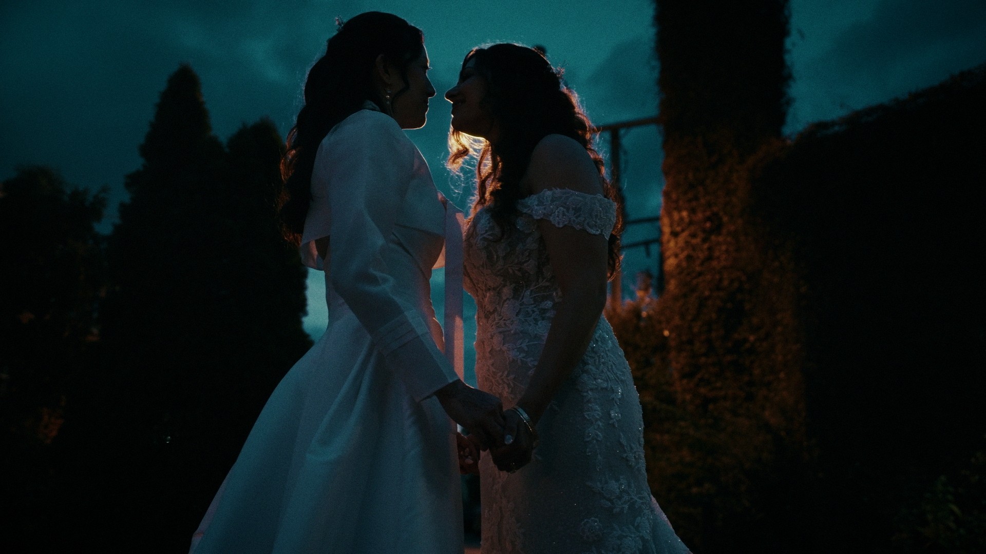 Two women in wedding dresses holding hands, facing each other outdoors at night with trees in the background.