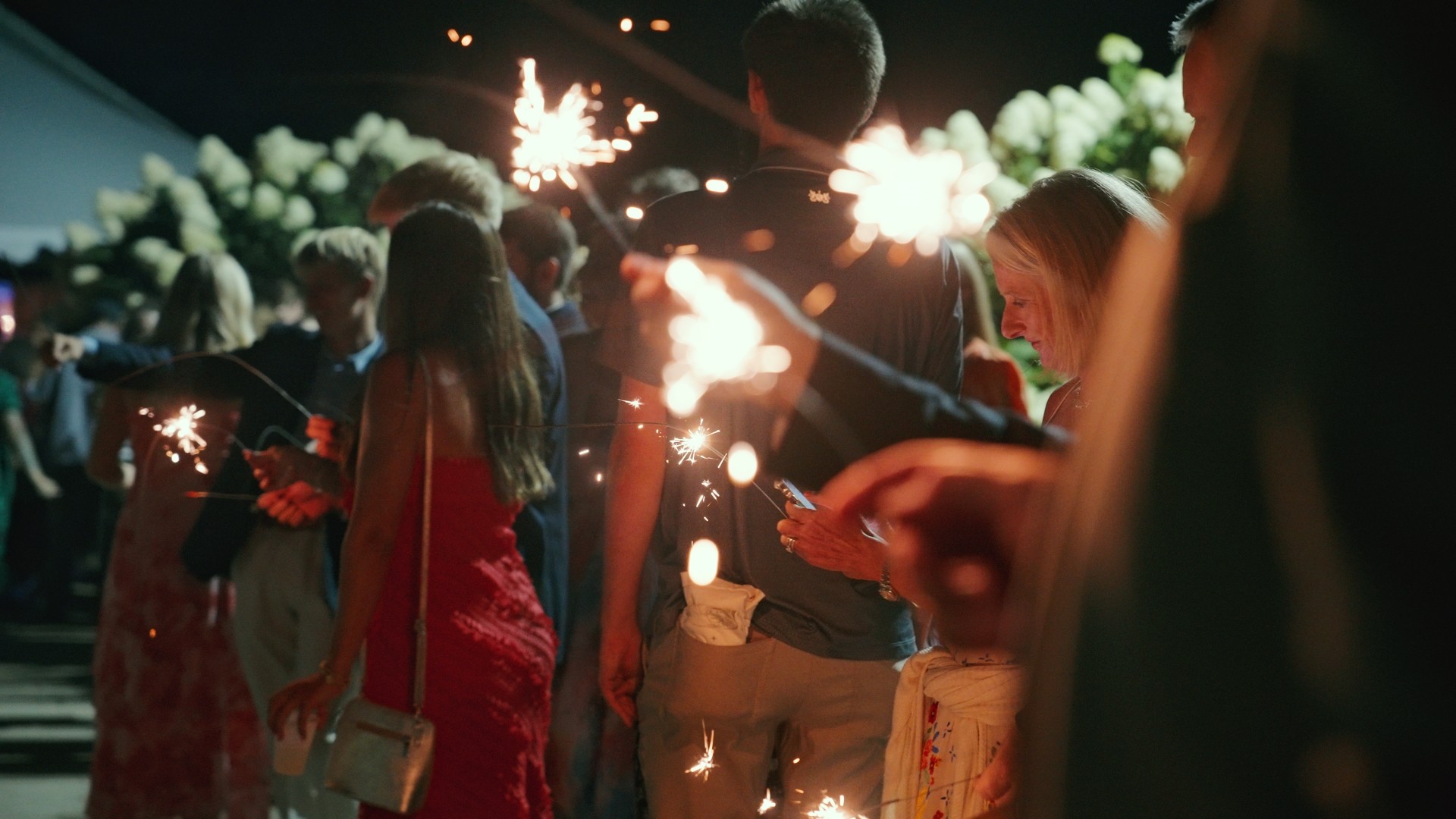 People holding sparklers at night, some wearing red dresses, with bright sparks and blurred background.