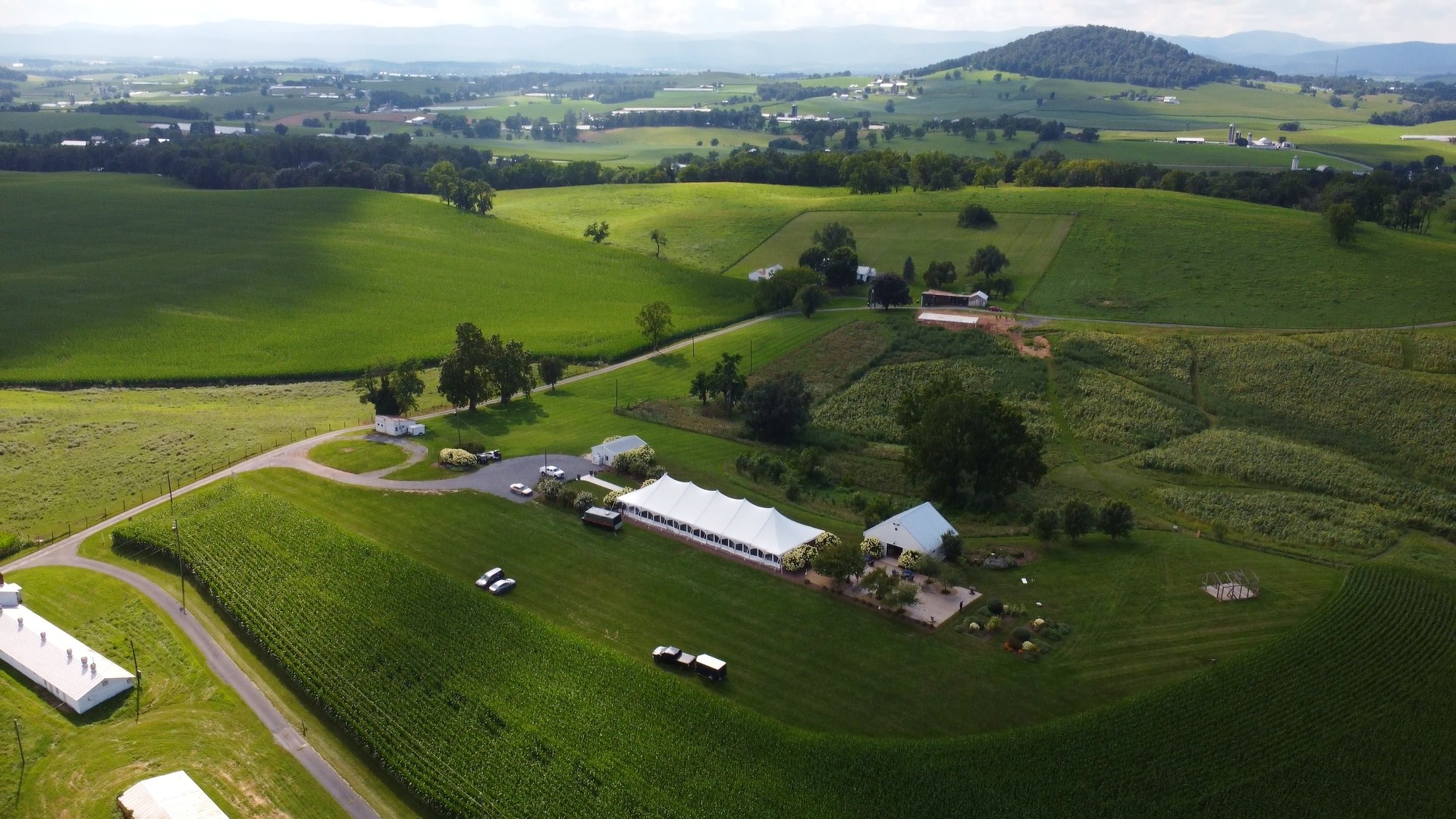 Aerial view of green fields, farm buildings, and roads in a rural landscape with hills in the background.
