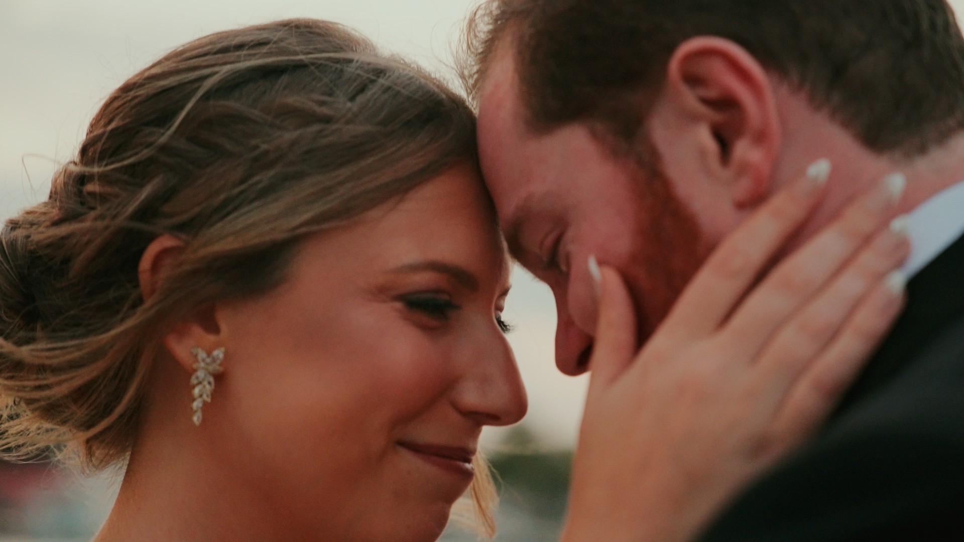 Close-up of a couple touching foreheads, smiling, with a blurred outdoor background.