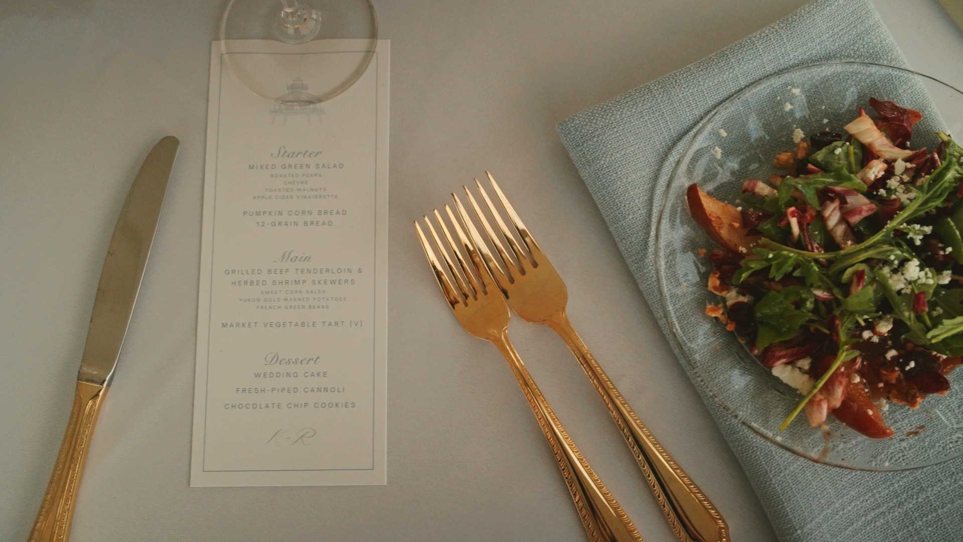 Table setting with knife, gold forks, a glass, a menu, and a salad on a blue napkin.