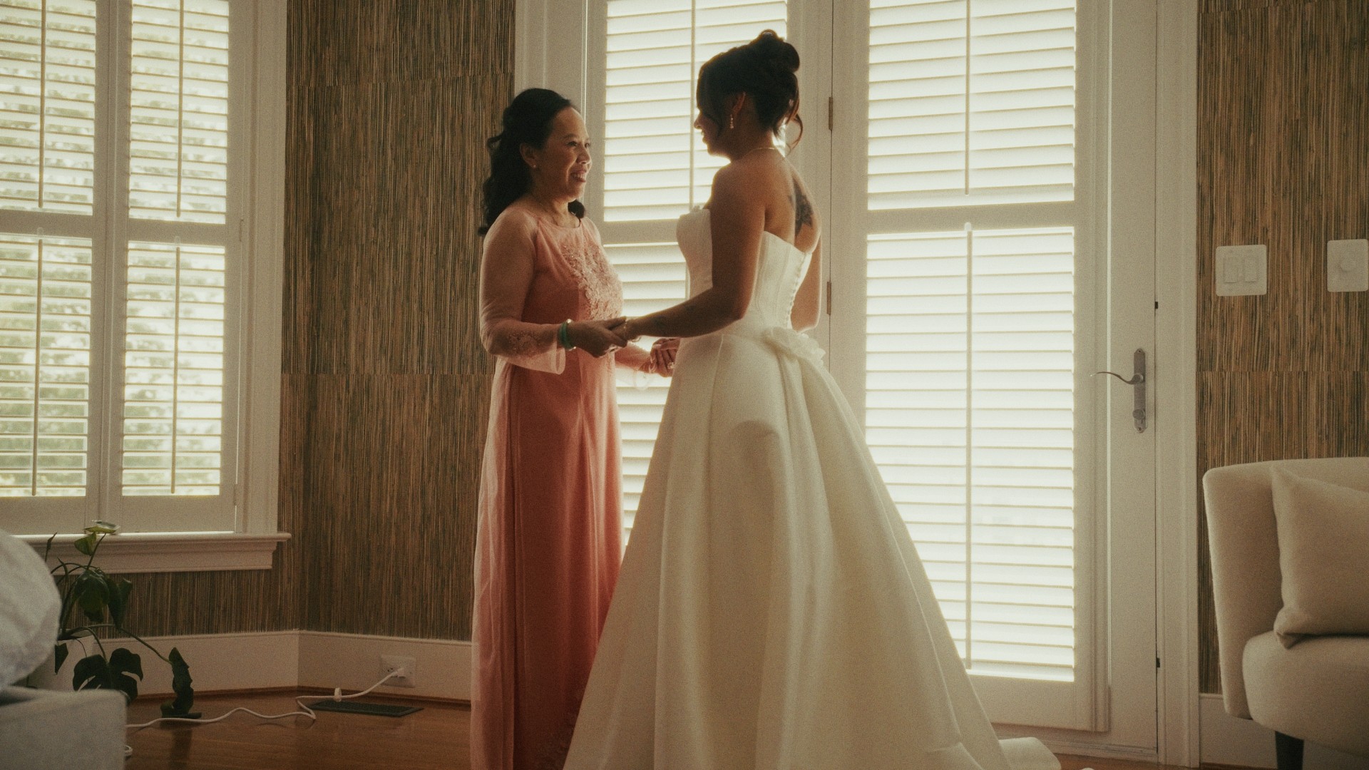 Two women stand indoors near windows with shutters, holding hands and facing each other, one in a pink dress and the other in a white gown.