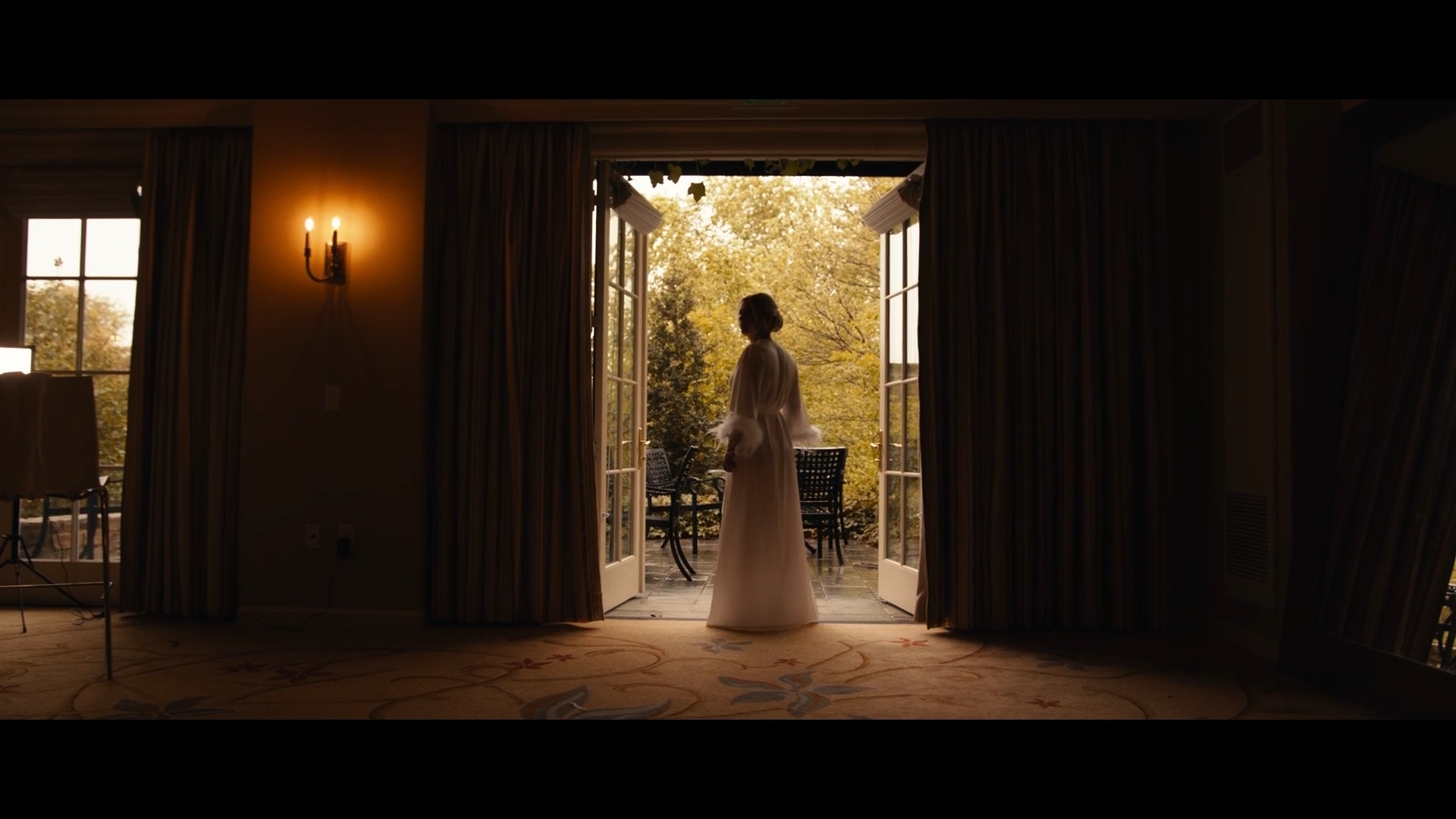 Person standing in doorway with open curtains, outdoor scenery visible, warm lighting inside, daytime.