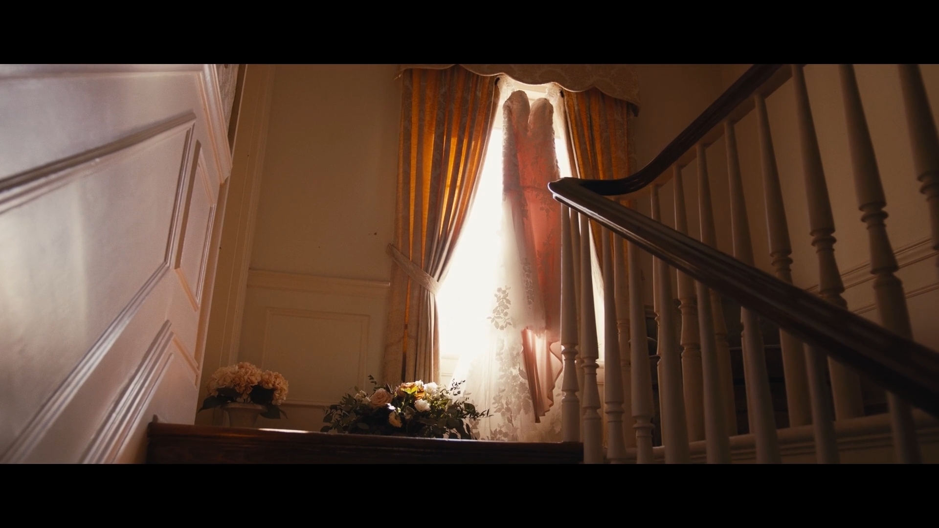 Sunlight streams through a window with curtains, illuminating a staircase and a flower arrangement on a table.