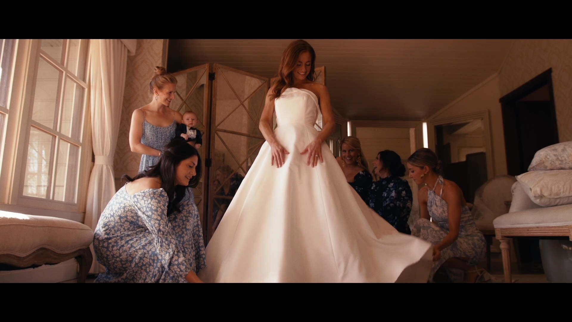 A bride in a white wedding dress stands surrounded by women in a room with natural light from a window.
