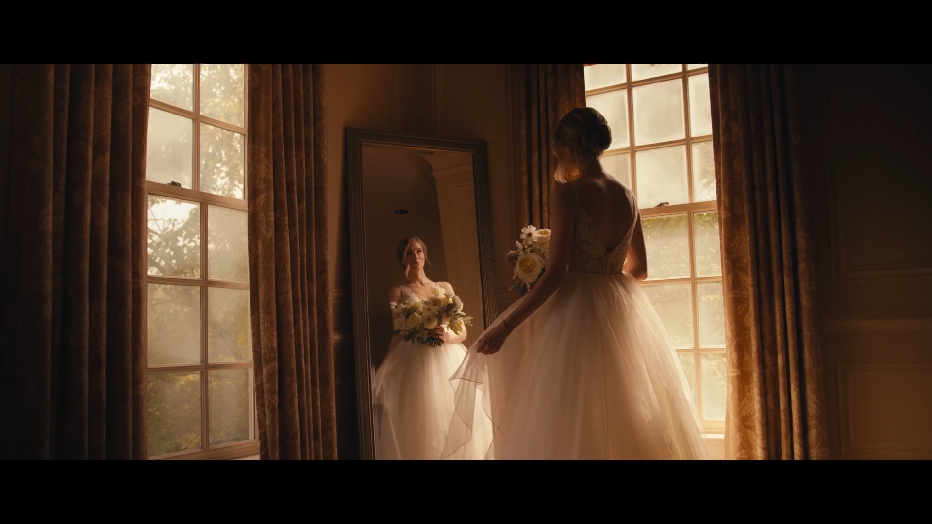 A woman in a wedding dress stands in front of a mirror, illuminated by sunlight through large windows, holding a bouquet.