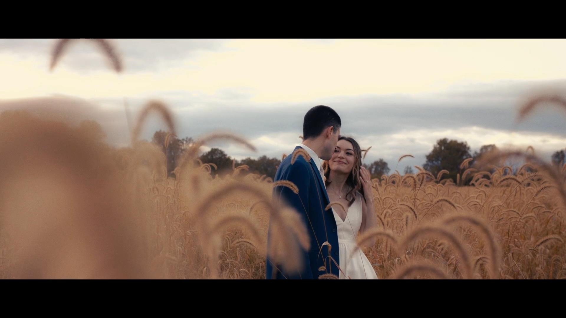 A couple stands in a field of tall grass, with trees and cloudy sky in the background.