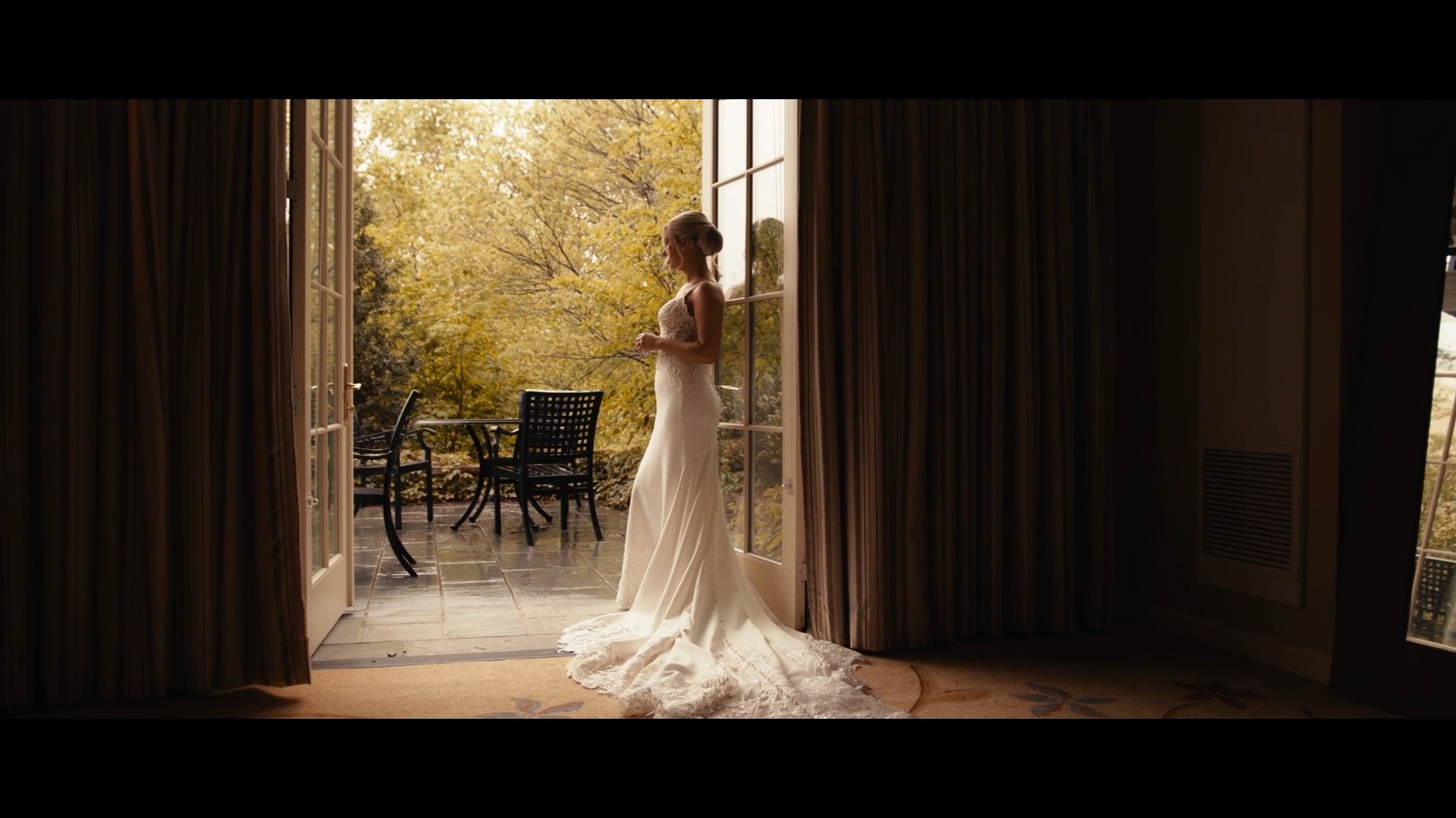 Woman in wedding dress standing in doorway opening to outdoor patio with trees, chairs, and table, sunlight filtering through trees.