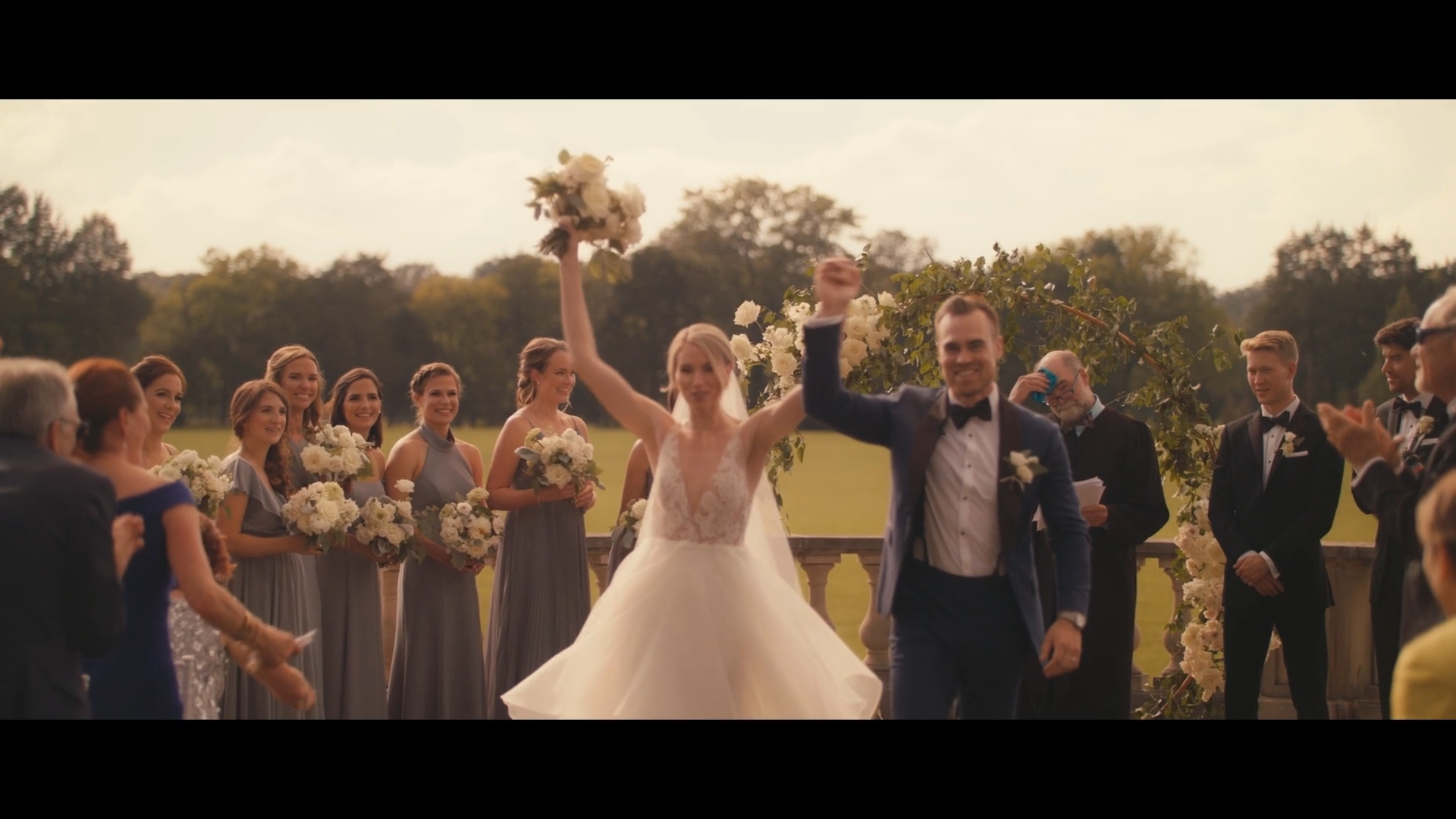 Bride and groom celebrating outdoors with friends and family, holding bouquets and glasses, trees in background.