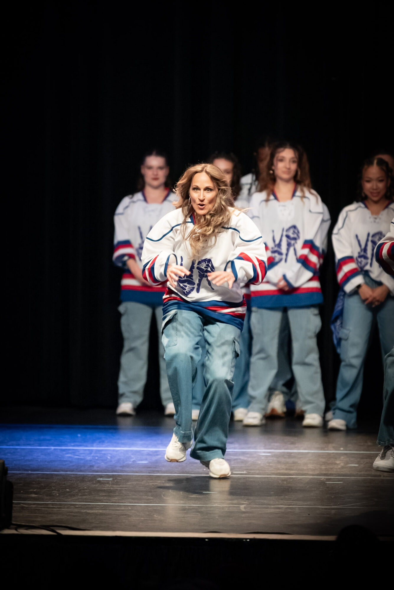 Group of young women performing on stage, one in front wearing a sports jacket and jeans, others in similar outfits behind.