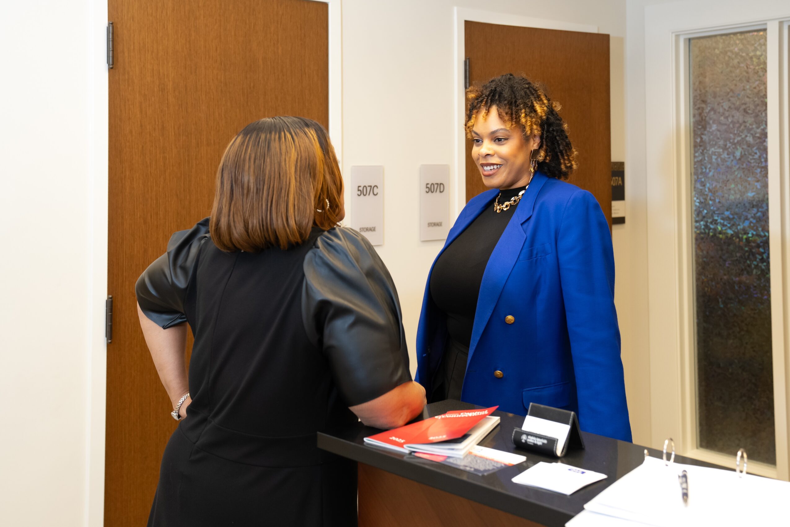 Two women talking in an office hallway, one in a black dress and the other in a blue blazer, smiling.