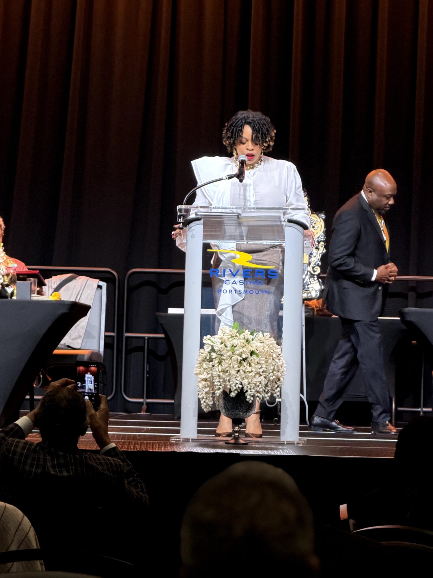 Woman speaking at a podium on stage with a black curtain background, a man in a suit nearby, audience in foreground.