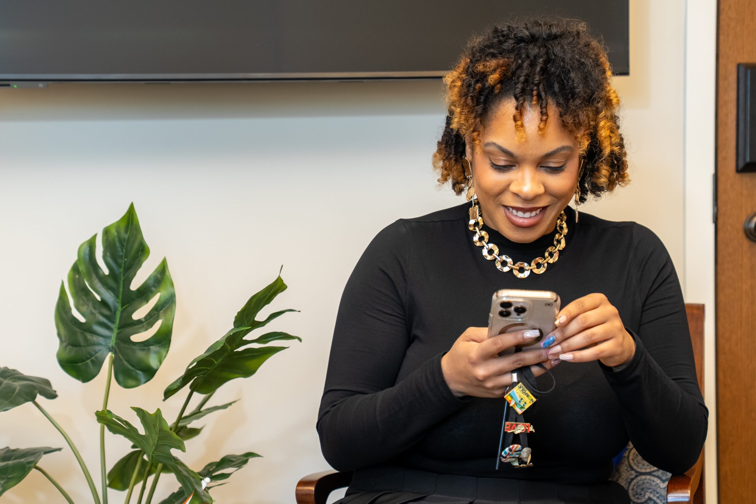 Woman with curly hair smiling while looking at her phone, sitting indoors with a large plant nearby.