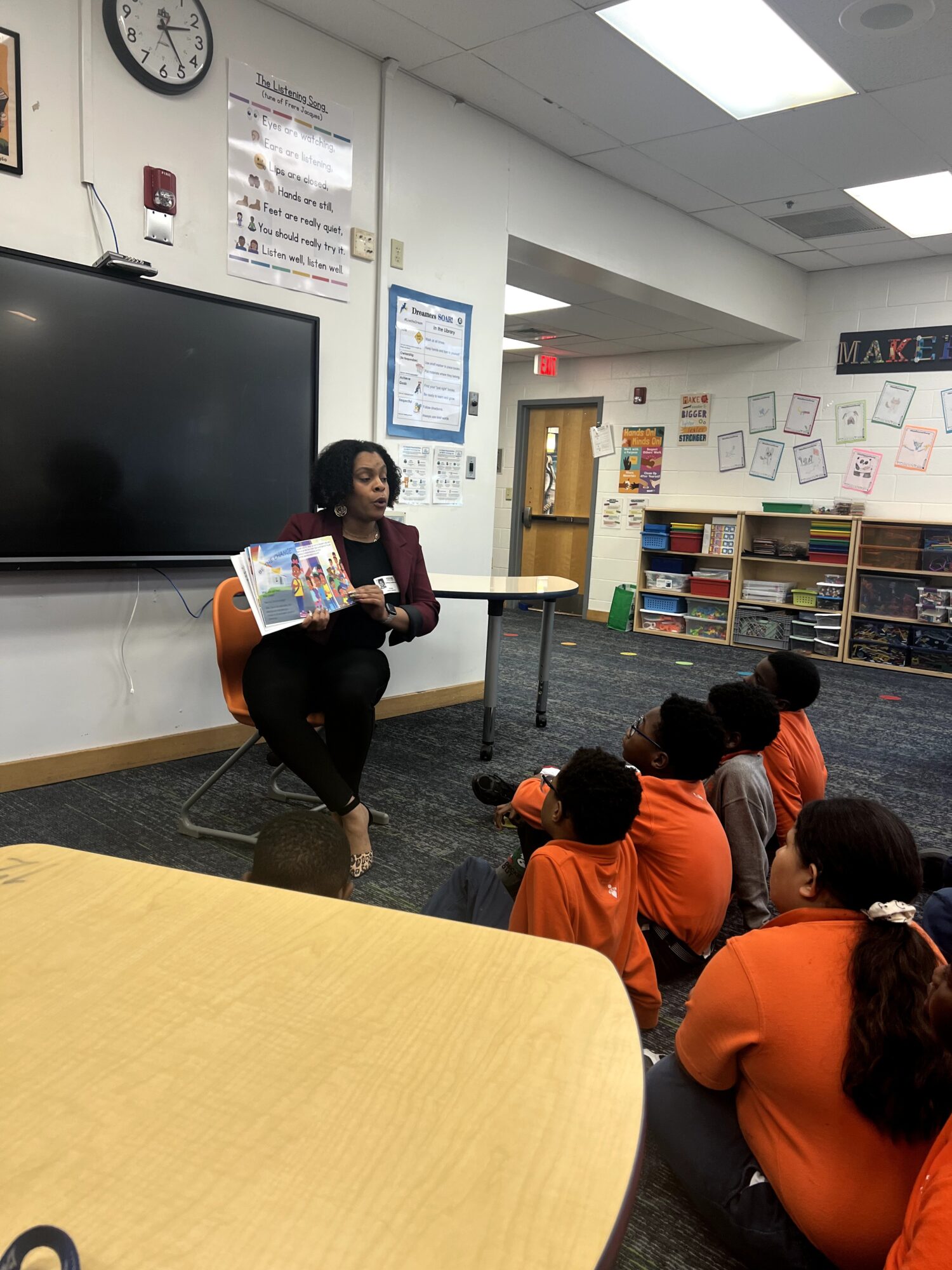 Teacher reading a book to students seated on the floor in a classroom with educational posters and shelves.