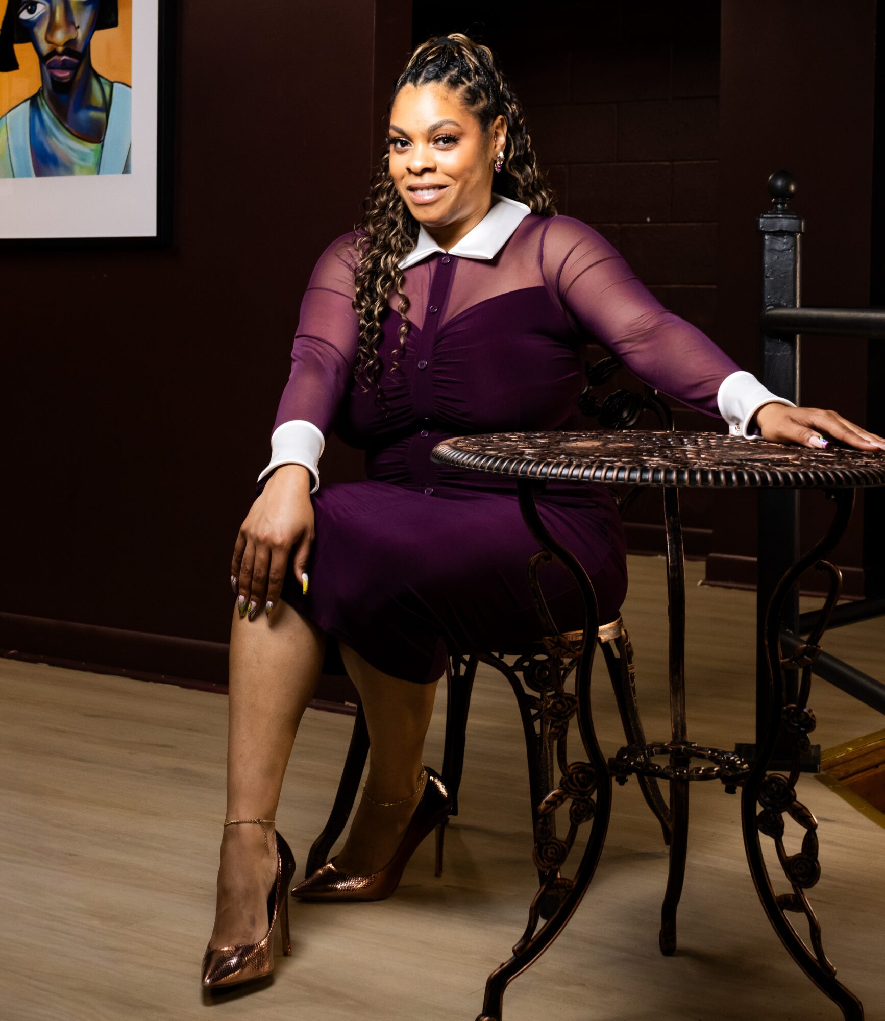 Woman sitting at a table with a dark background, wearing a purple dress with sheer sleeves, smiling, with artwork on the wall.