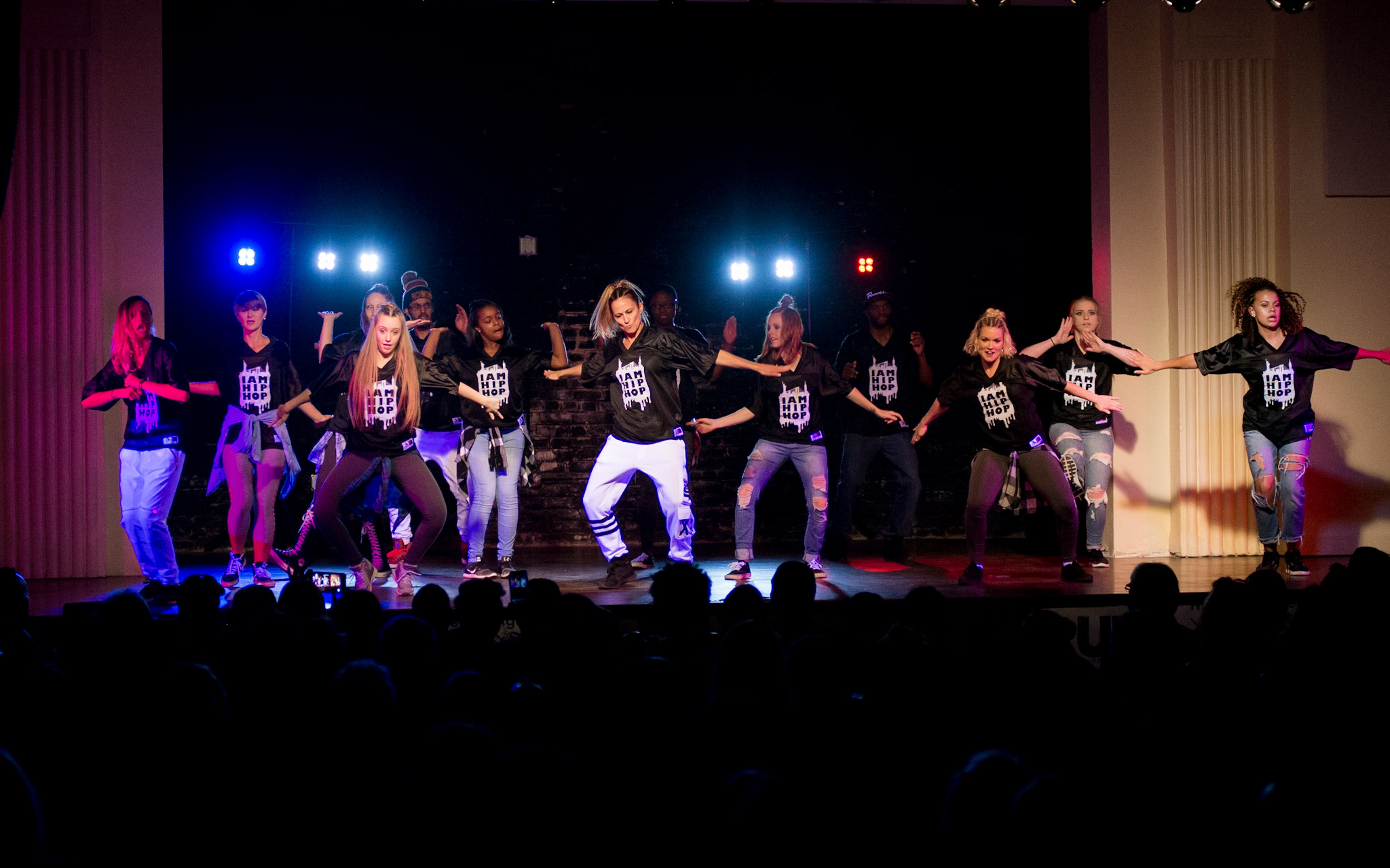 Group of dancers performing on stage with colorful lights, audience in foreground, stage curtains on sides.