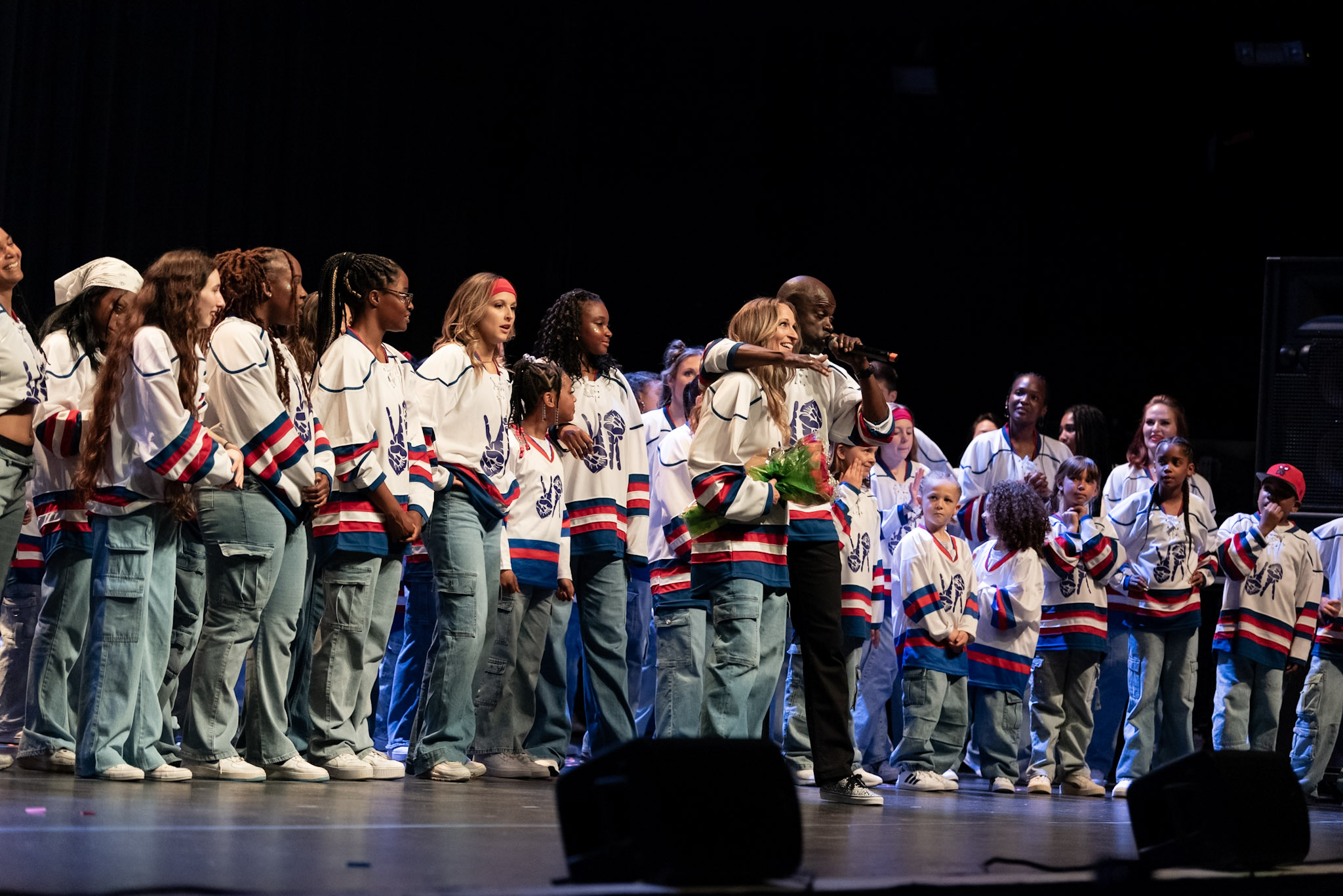 Group of people on stage wearing matching jerseys, some hugging, with a dark background.