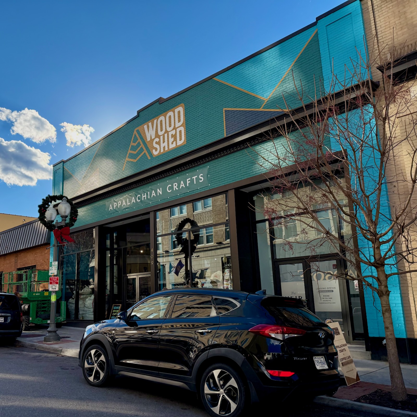 Storefront with signage for Wood Shed and Appalachian Crafts, black car parked outside, leafless tree, blue sky with clouds.