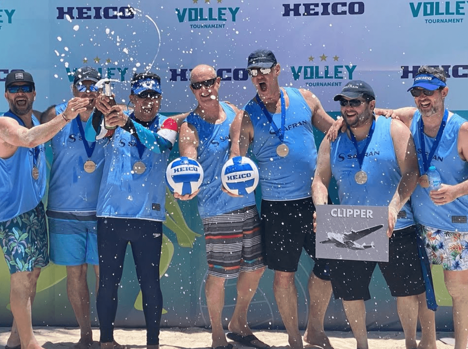 Group of seven people celebrating with medals and a trophy on a beach, holding a box labeled 'CLIPPER', smiling and spraying water.
