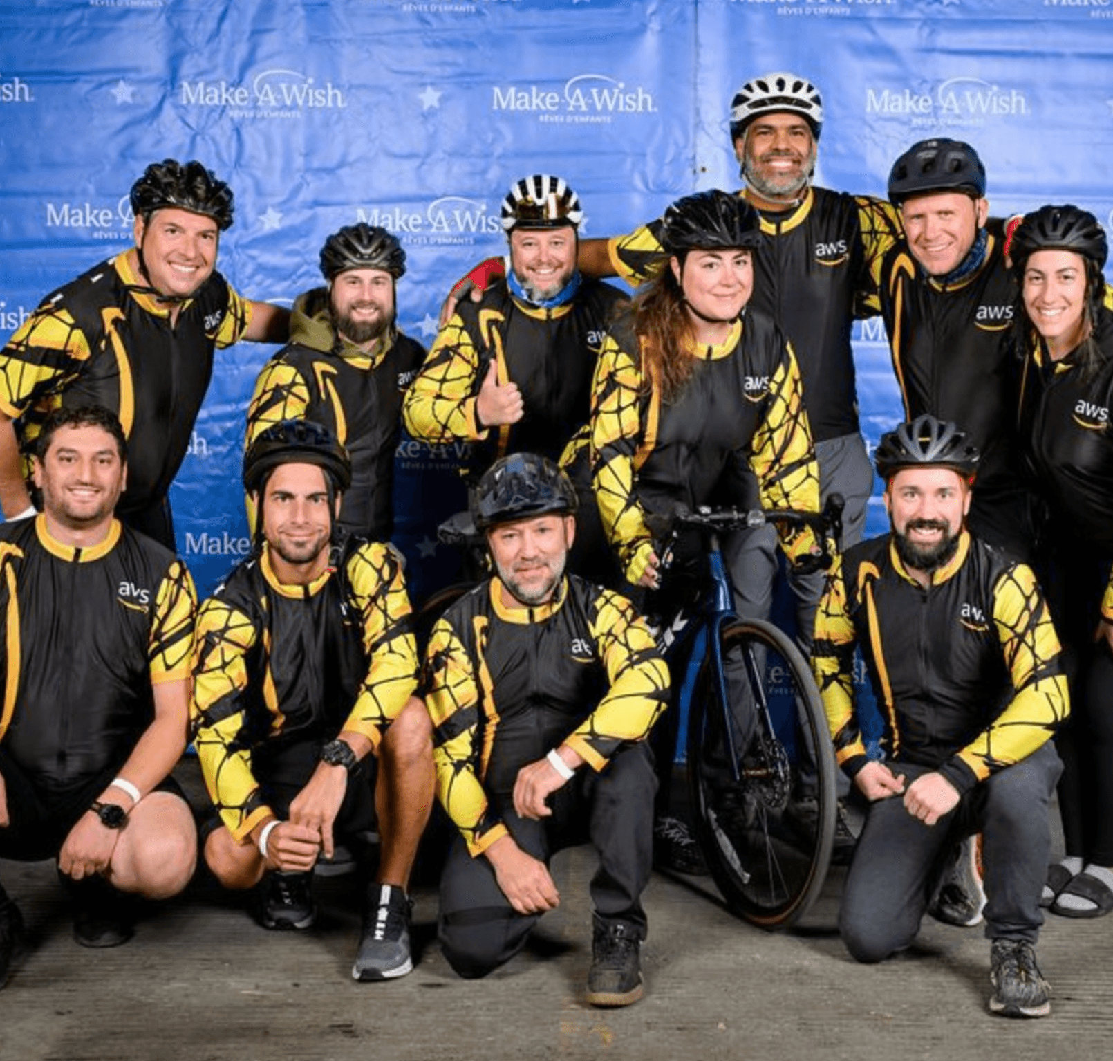 Group of ten people in matching black and yellow jackets, some wearing helmets, posing with a bicycle in front of a blue backdrop.
