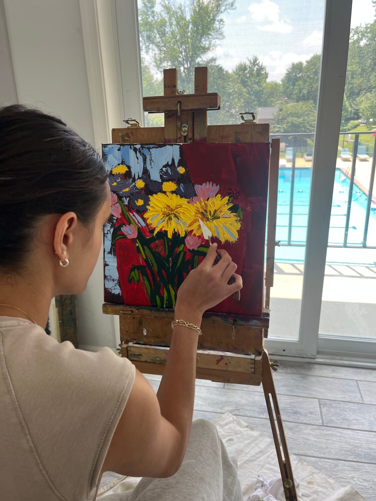 Woman painting a colorful floral artwork on an easel indoors, with a window showing trees outside.