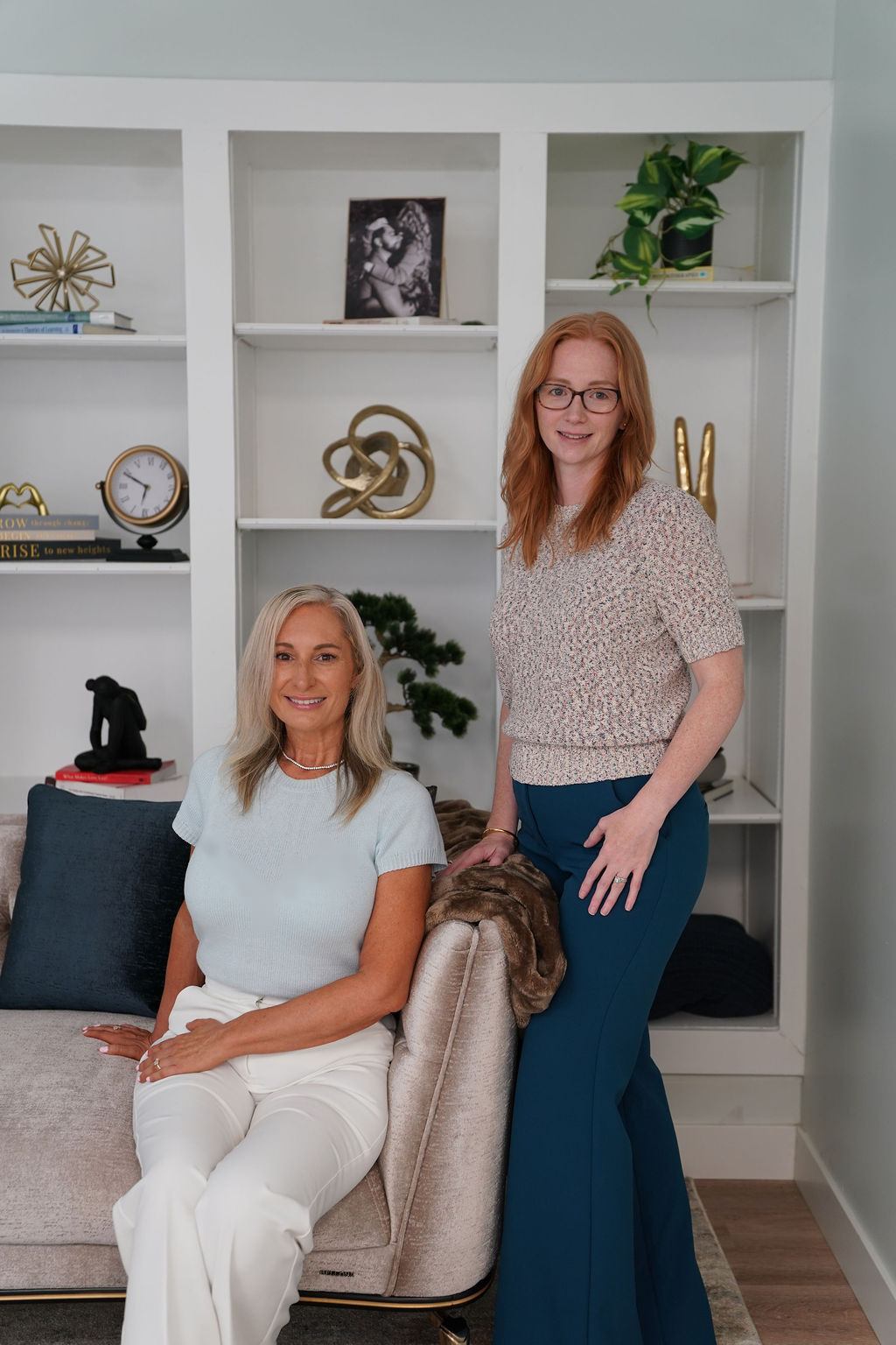 Two women in a living room with white shelves, decorative items, and a beige sofa, one sitting and one standing, smiling.