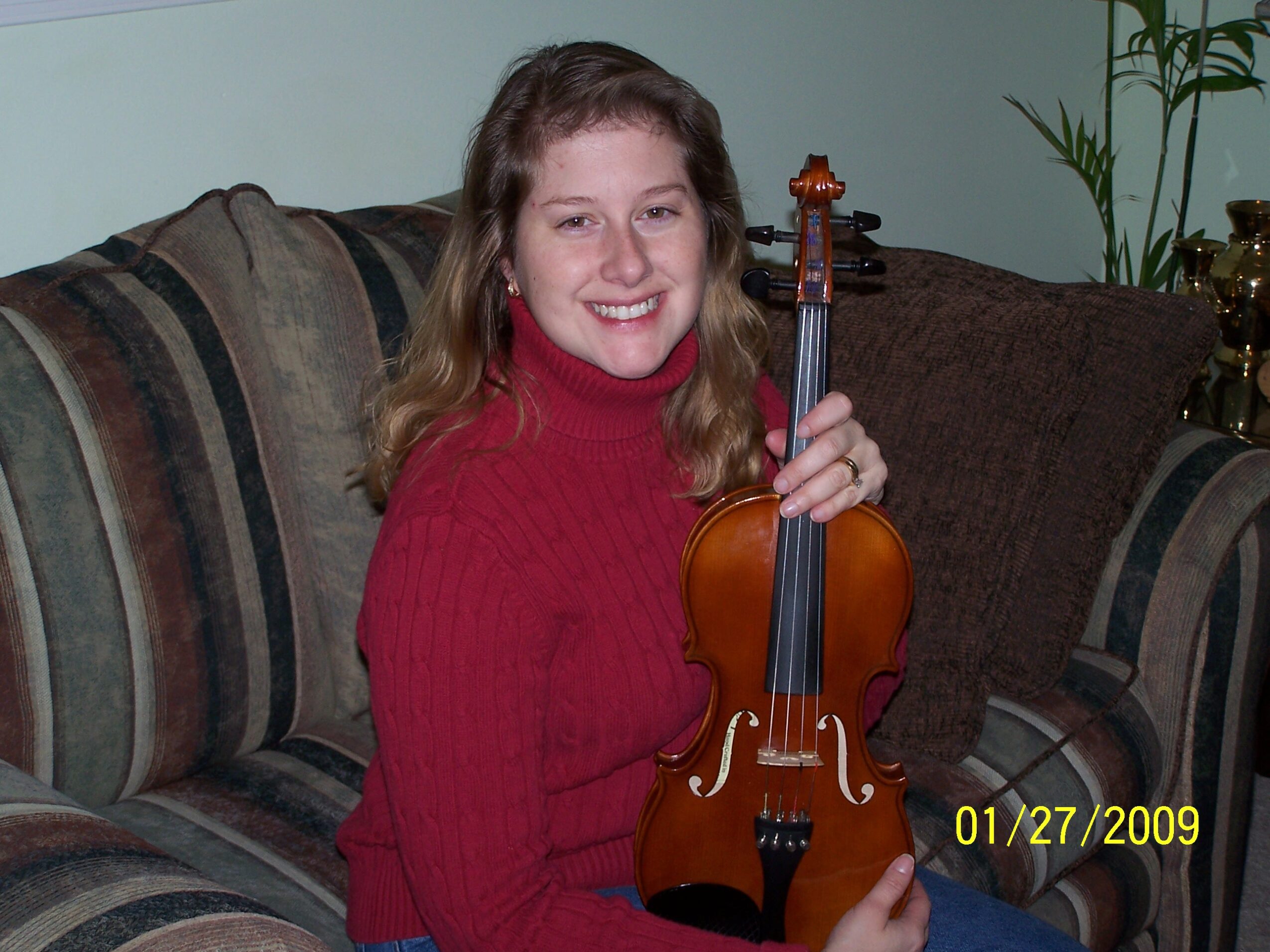 Young woman with long hair smiling, holding a violin, sitting on a striped sofa in a living room.